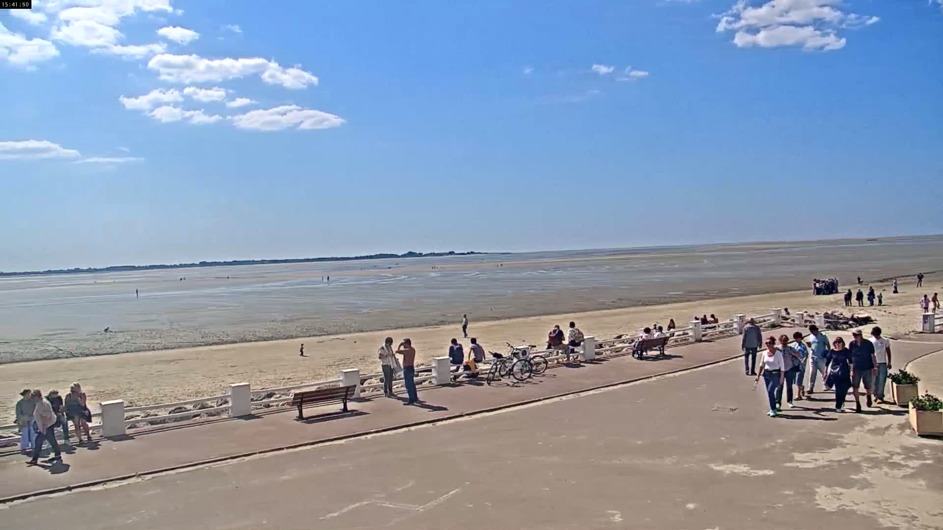 A paved walkway overlooks a wide expanse of exposed sand and mudflats at low tide, with people walking and sitting along the promenade under a mostly sunny sky with scattered clouds.