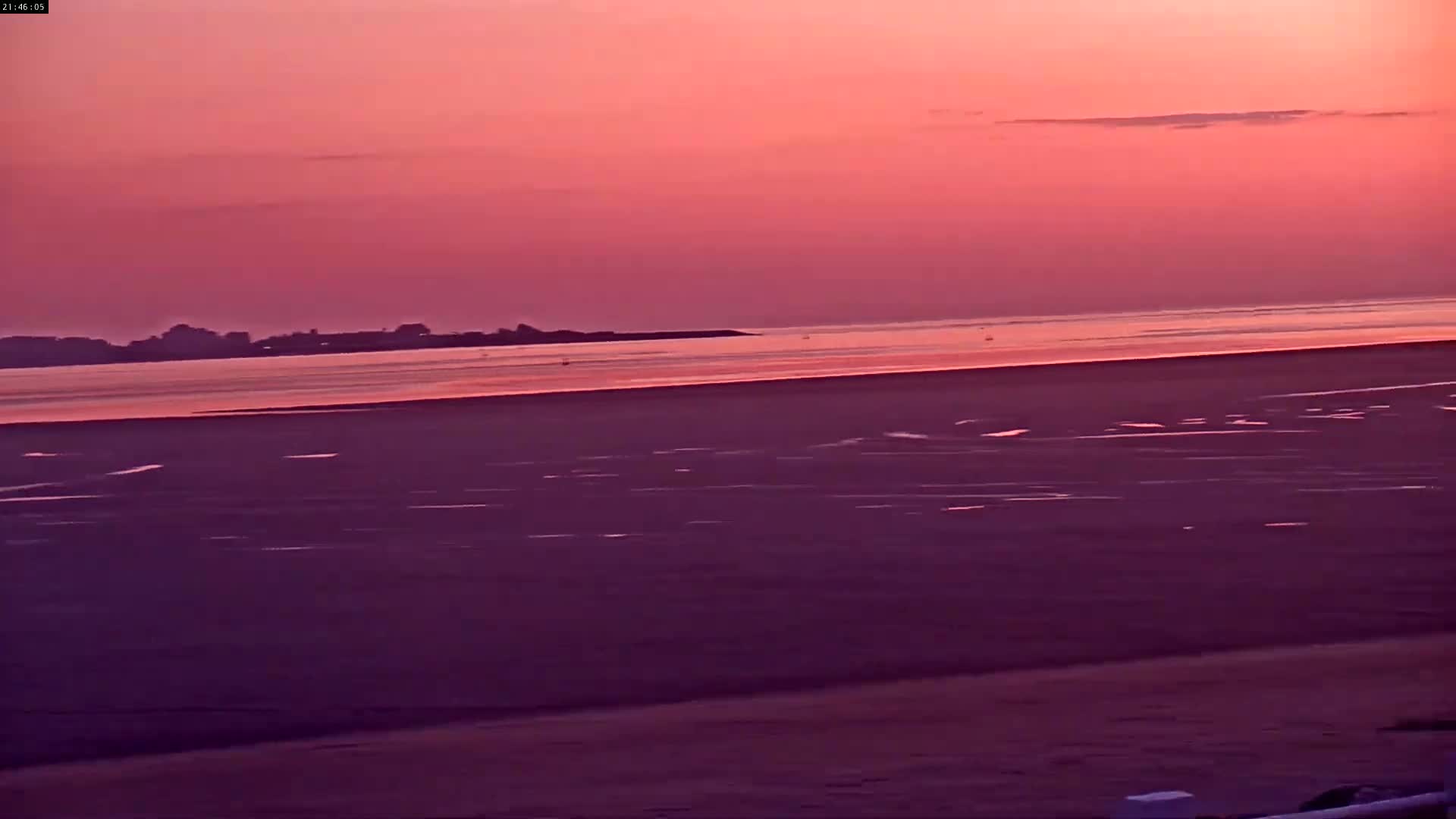 A pink sunset illuminates a calm body of water and a flat sandy beach.