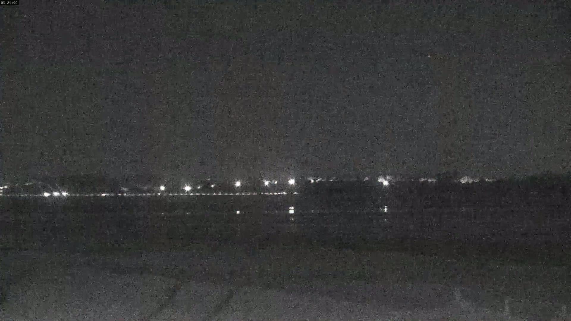 A nighttime view shows a line of distant lights across a dark body of water under a clear, dark sky.