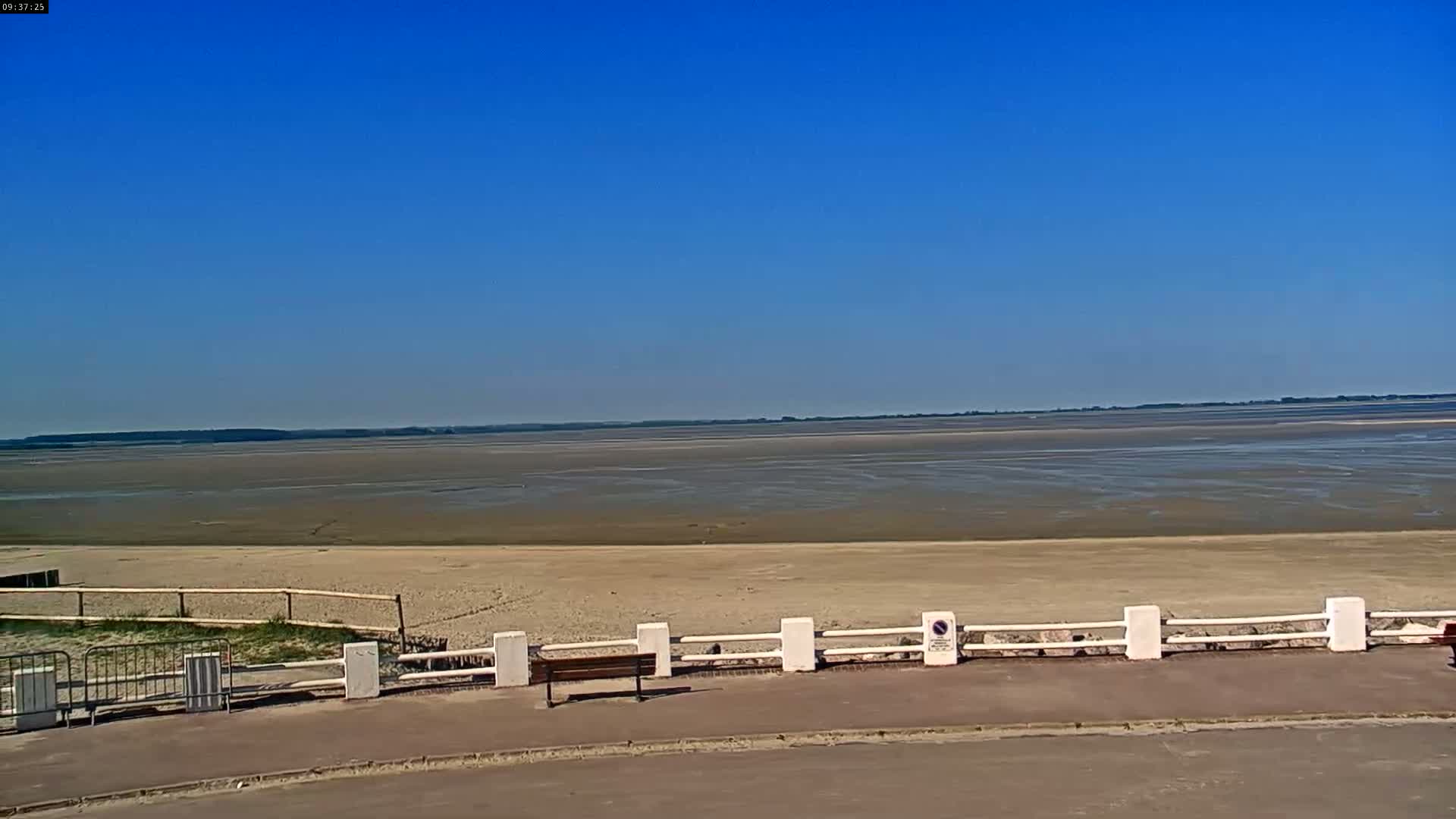 A mostly sunny day reveals a wide expanse of light brown, sandy beach and calm, shallow water extending to a distant shoreline under a clear blue sky.