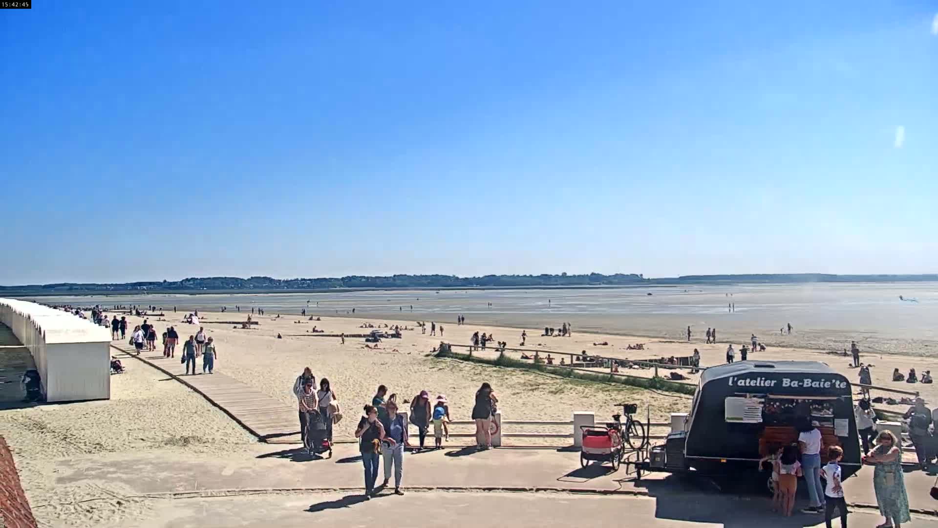 A sunny day at a beach shows many people sunbathing and walking along the sand, with a food stand near a boardwalk and rows of beach huts.