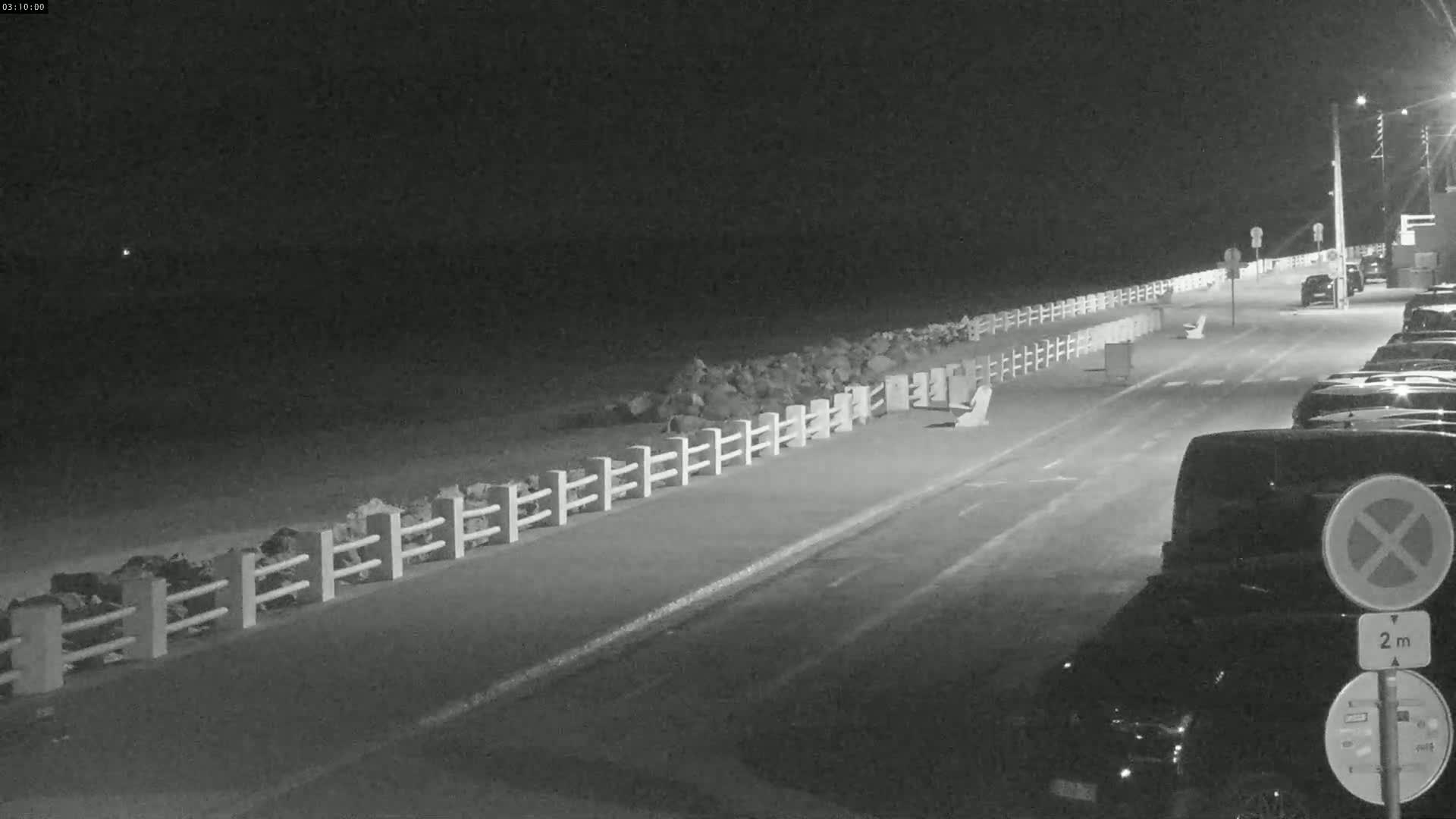 A nighttime, black and white view shows a paved road next to a rocky shoreline with a white fence, and several cars parked along the roadside.