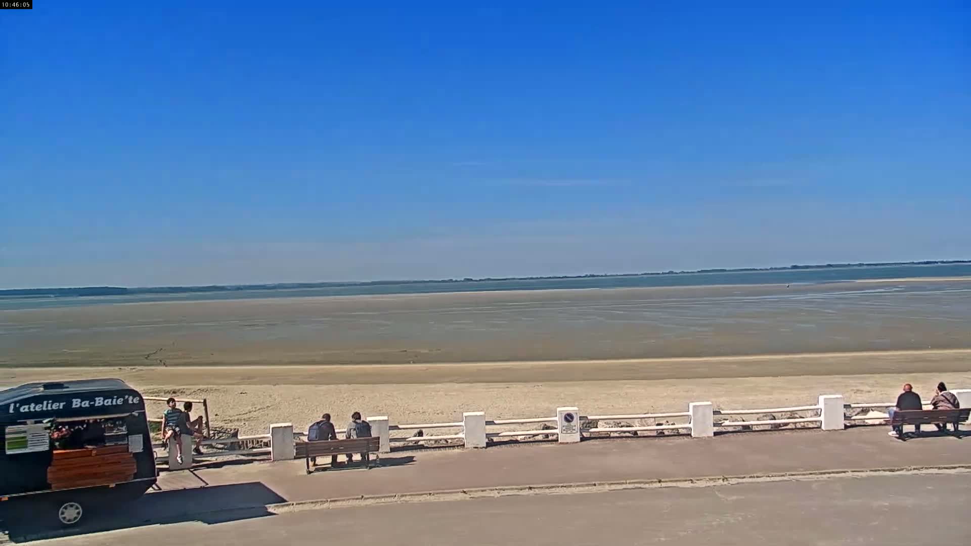 A wide expanse of exposed sand and shallow water stretches to a distant shoreline under a clear blue sky, with several people sitting on benches near a food trailer.