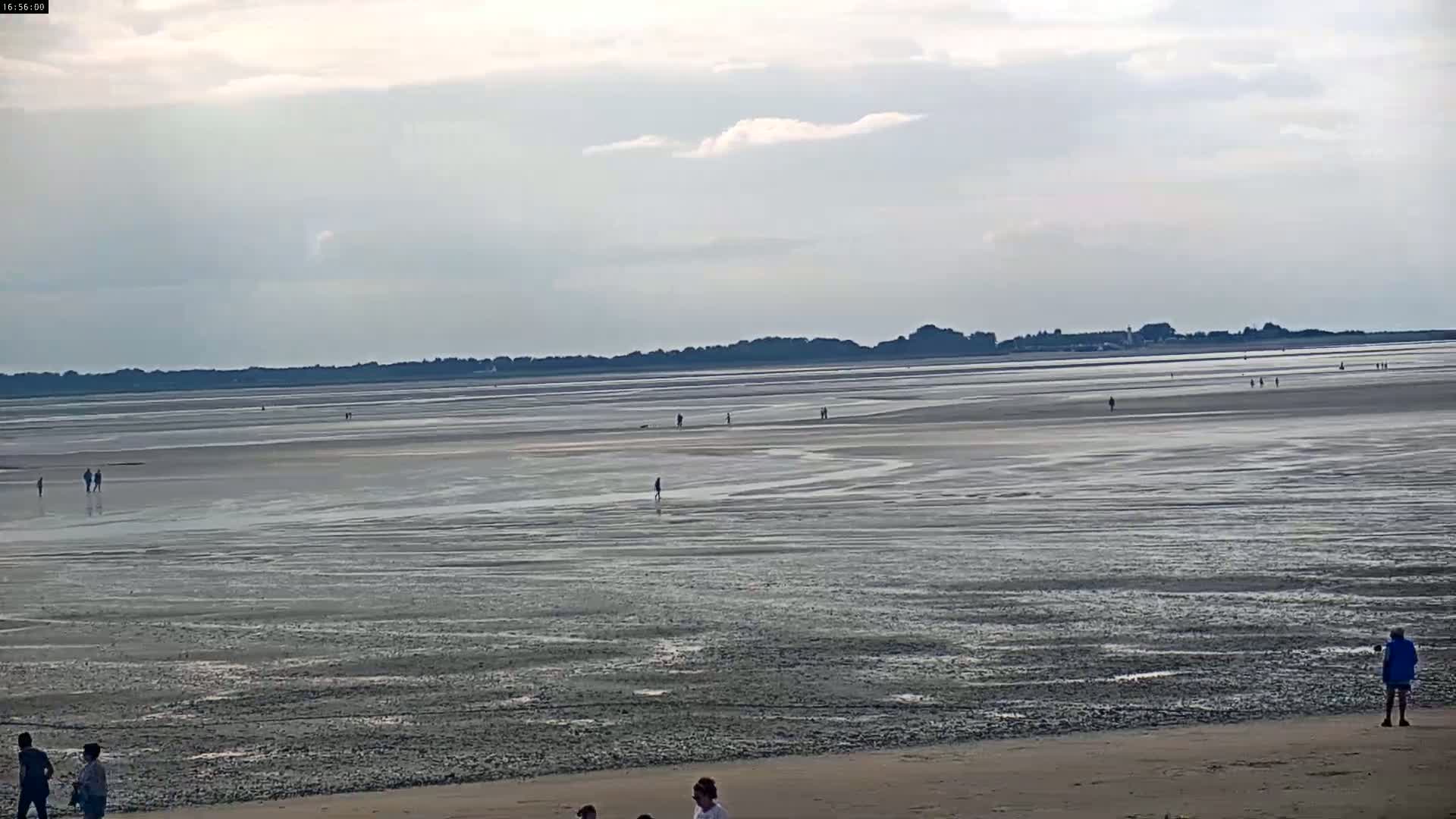 A wide expanse of exposed, mud-flat seabed stretches to a distant shoreline under a cloudy sky, with scattered people walking on the mud and a few more standing on a sandy beach in the foreground.