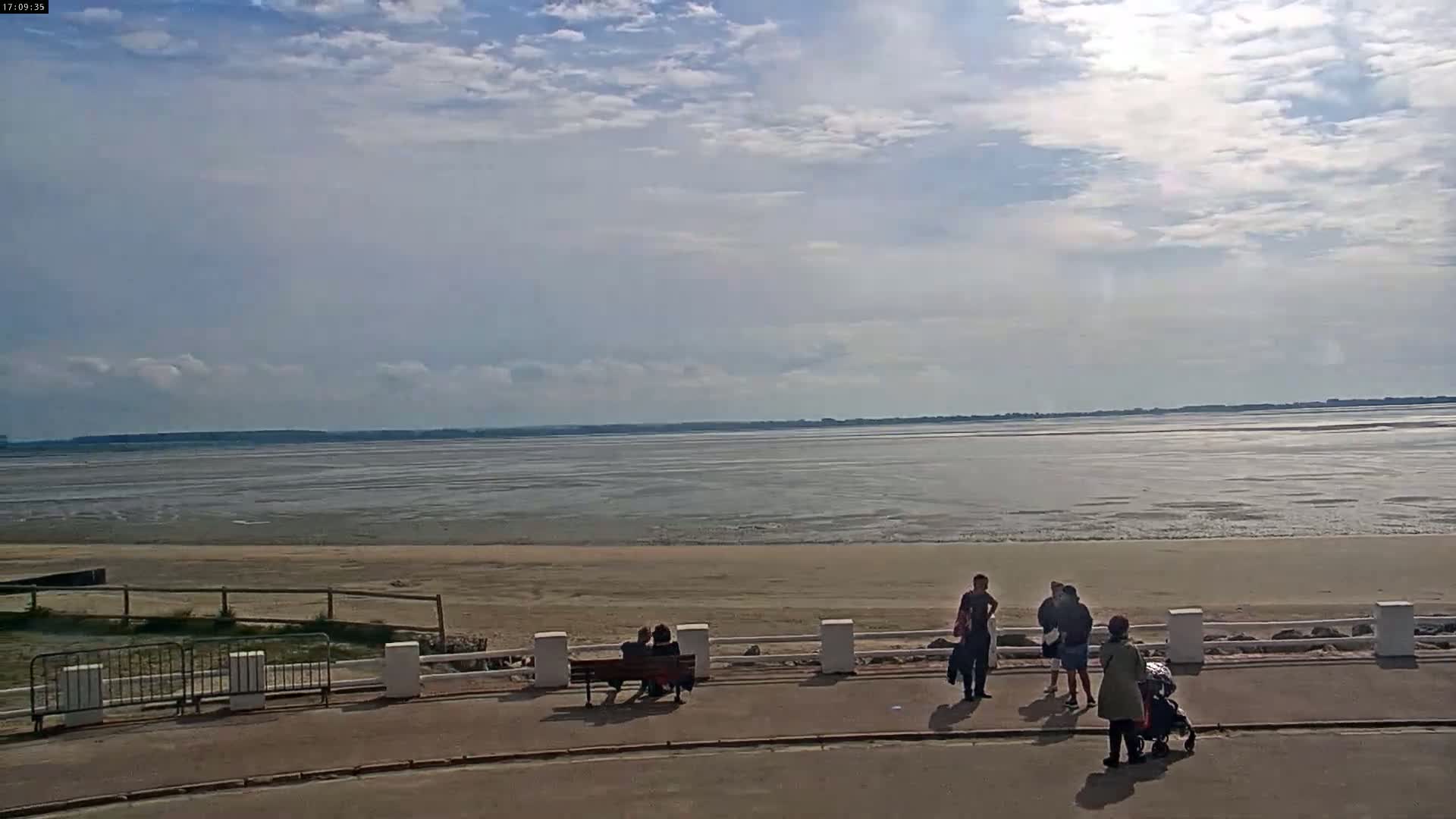 A partly cloudy day reveals a wide expanse of calm, shallow water receding to reveal a sandy beach, where several people stroll and sit on a bench near a low white railing.