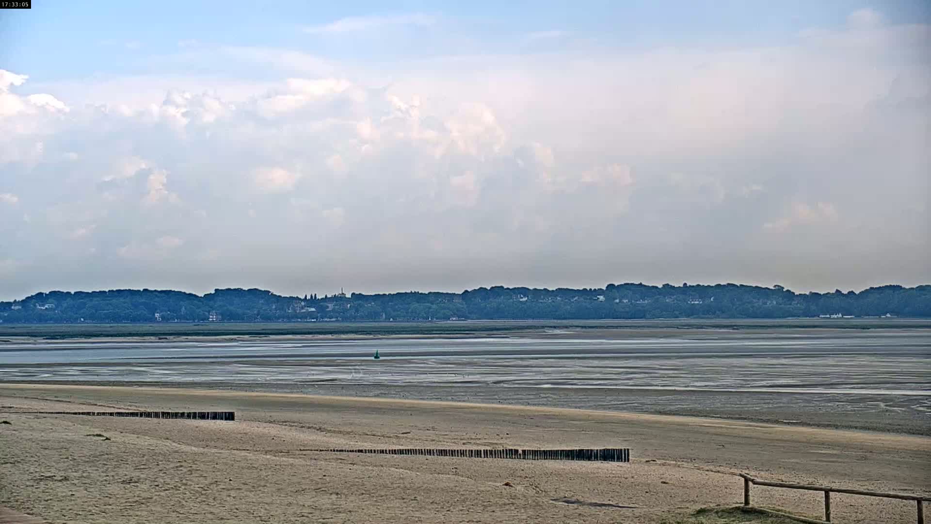A sandy beach stretches toward a wide expanse of exposed mudflats and water, under a partly cloudy sky, with a distant line of trees and buildings visible on the horizon.