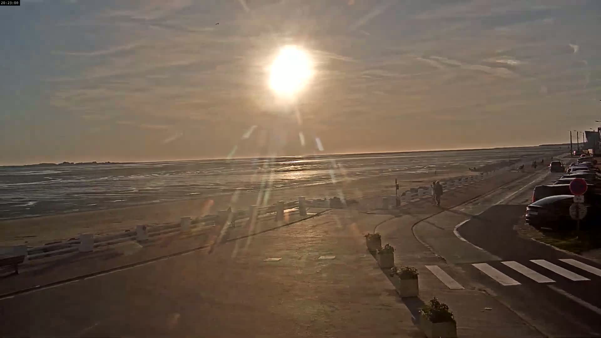 A low-lying sun shines over a calm, wide expanse of water and sandy beach, with a paved road and parked cars visible along the shore.