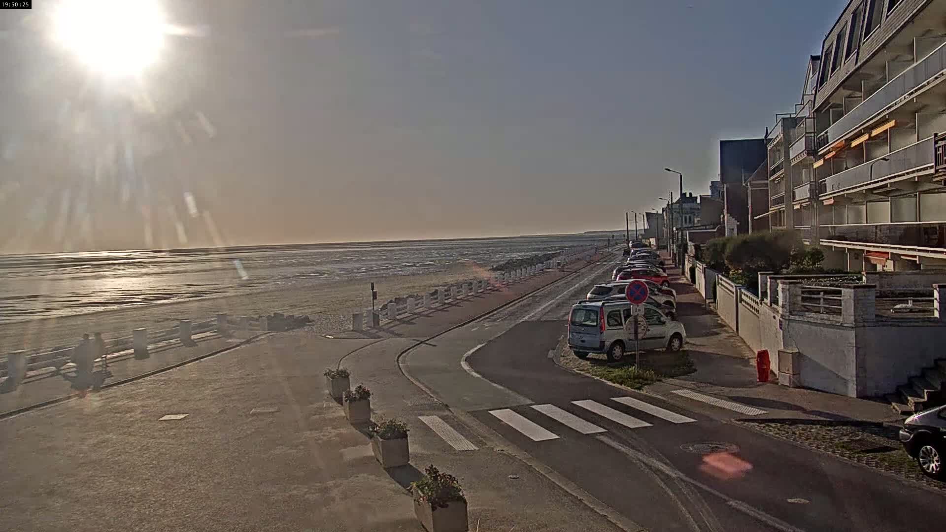 A sunny day at the beach shows a road leading to the ocean, cars parked along the street next to apartment buildings, and a pedestrian crossing.