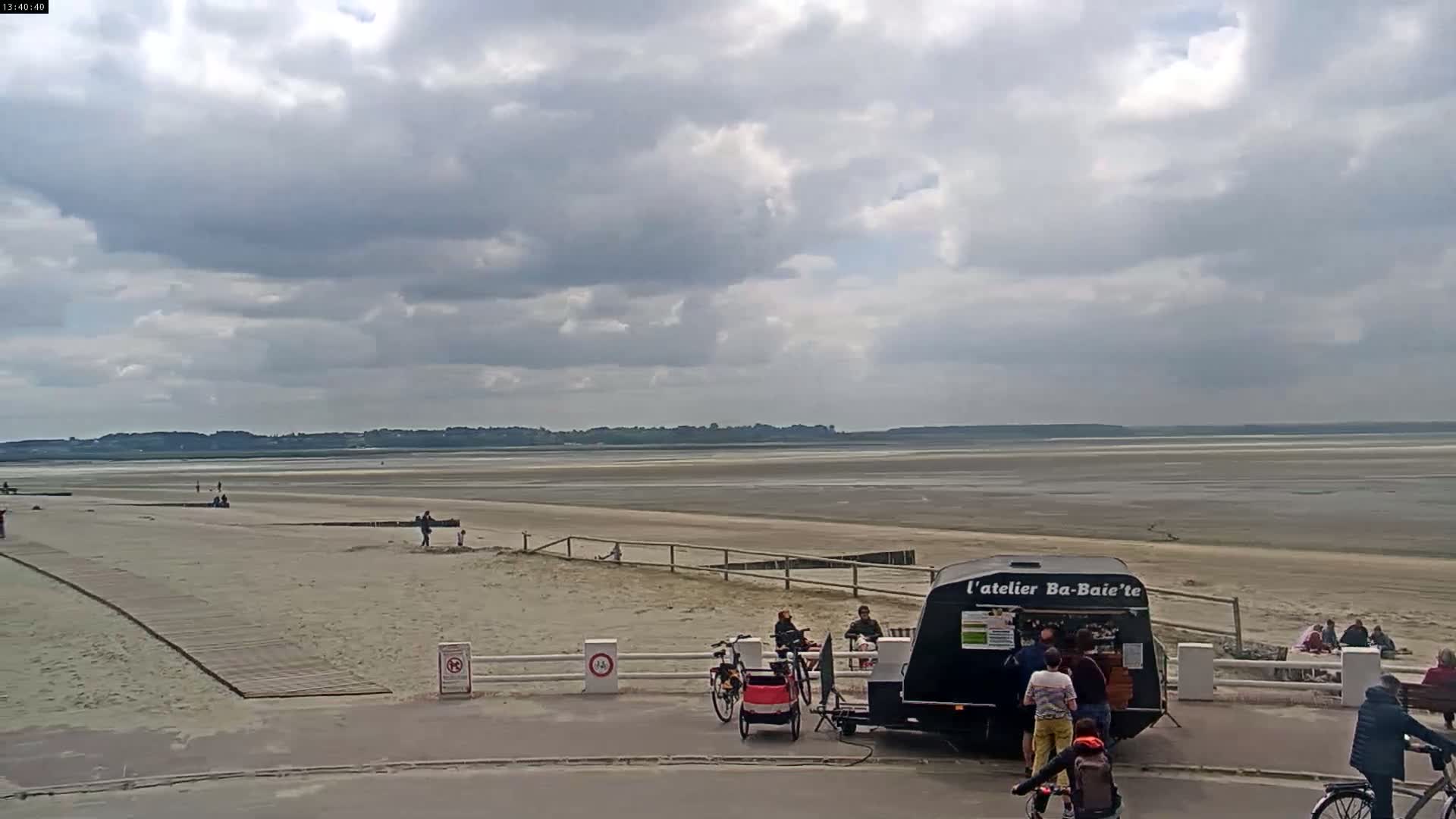 A sandy beach stretches to a calm, wide expanse of shallow water under a mostly cloudy sky, with several people near a food trailer and bicycles parked nearby.