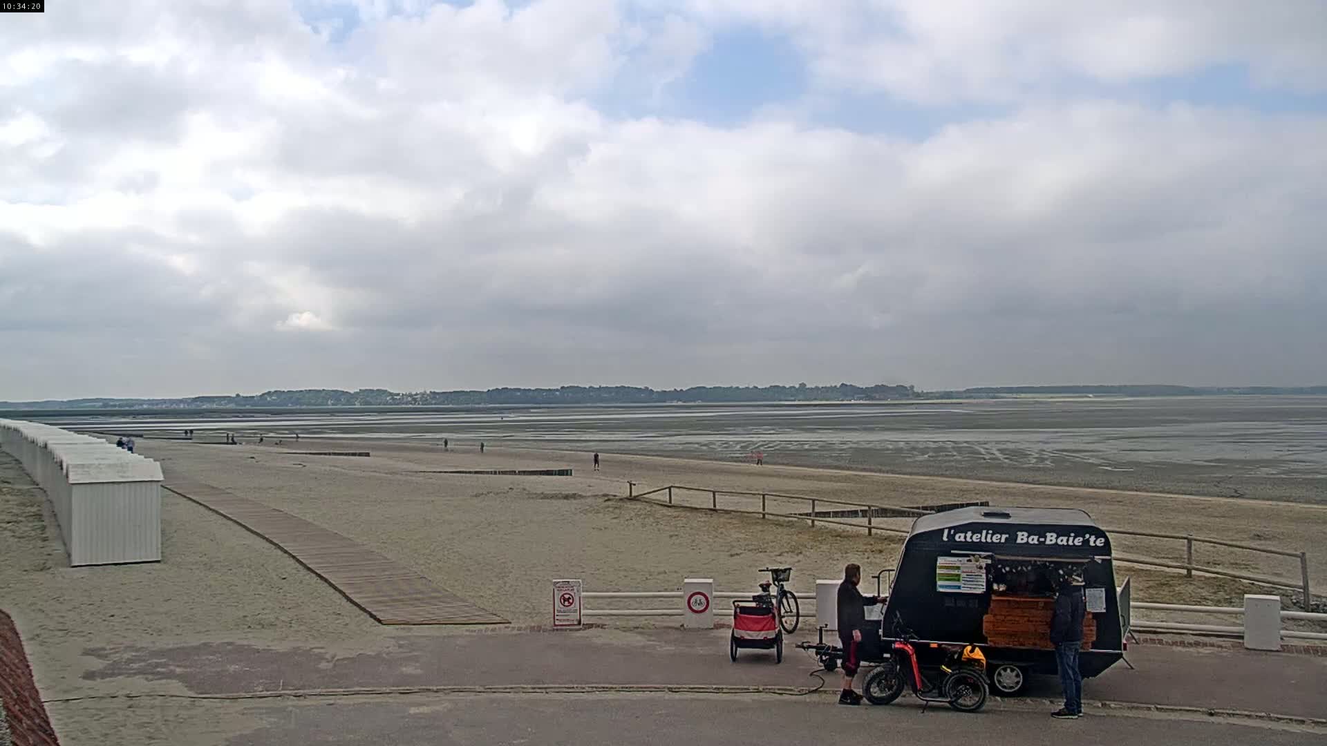 A sandy beach stretches to a wide expanse of mudflats under a cloudy sky, with a small food cart and several people near a boardwalk.