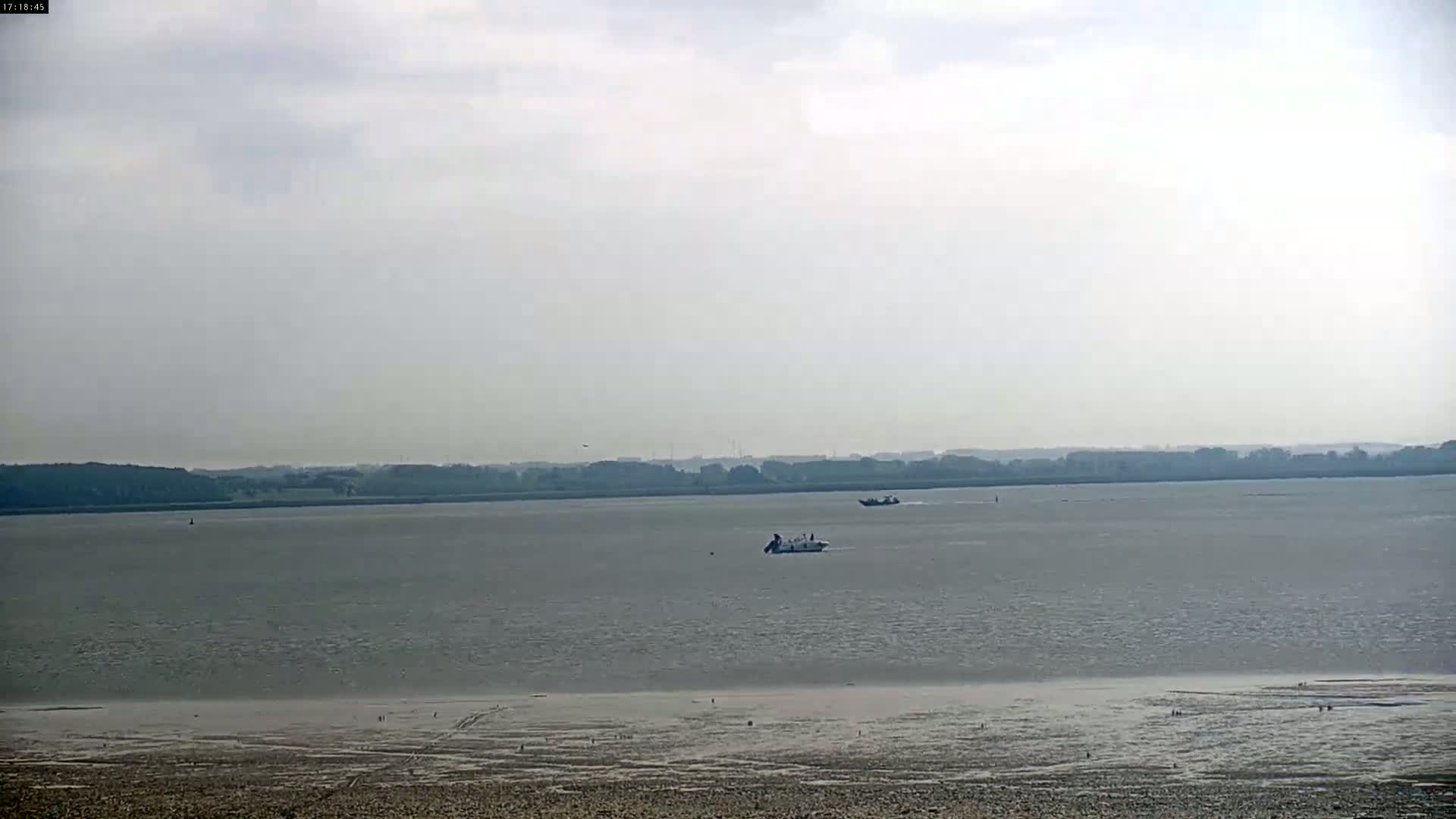 A wide, calm body of water stretches to a distant shoreline under a cloudy sky, with a few small boats visible on the water and a muddy, exposed shore in the foreground.
