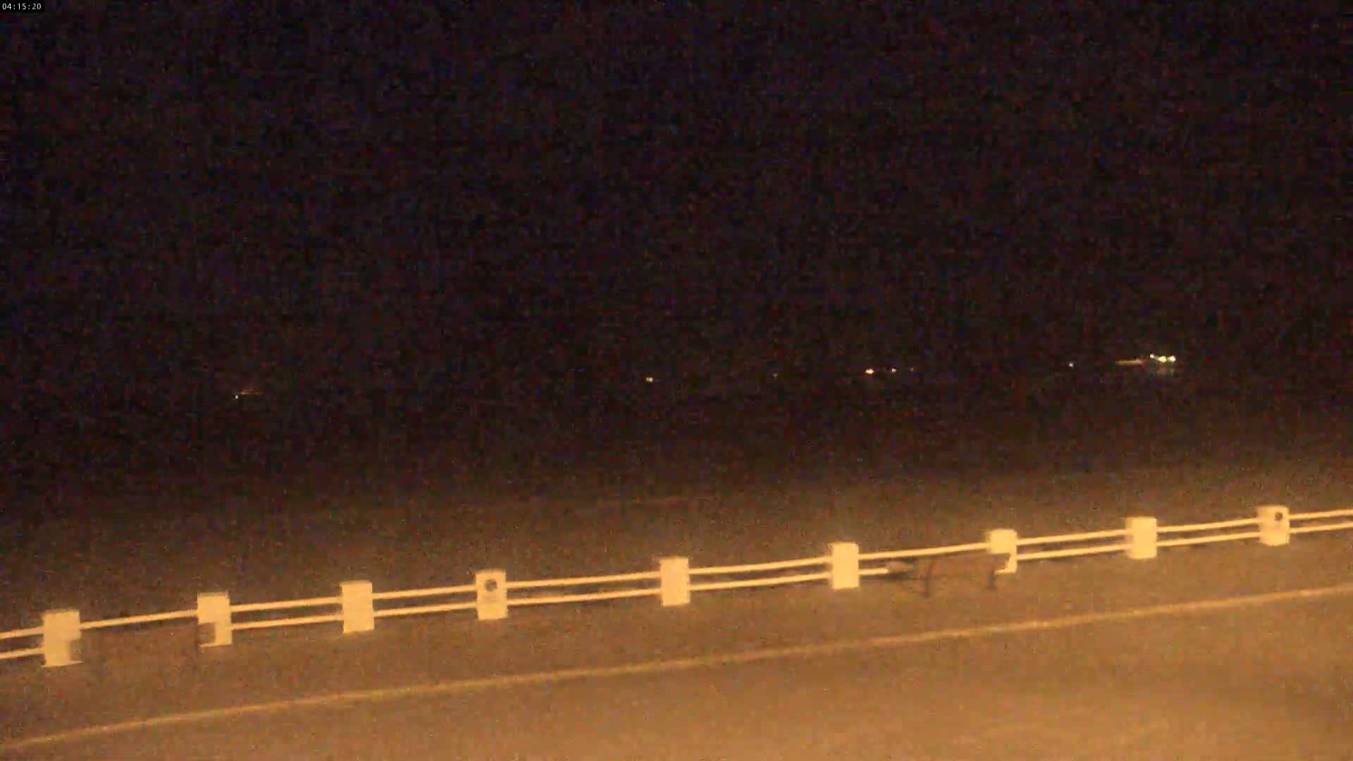 A nighttime view of a road with a low white fence, showing faint lights in the distance under a dark sky.