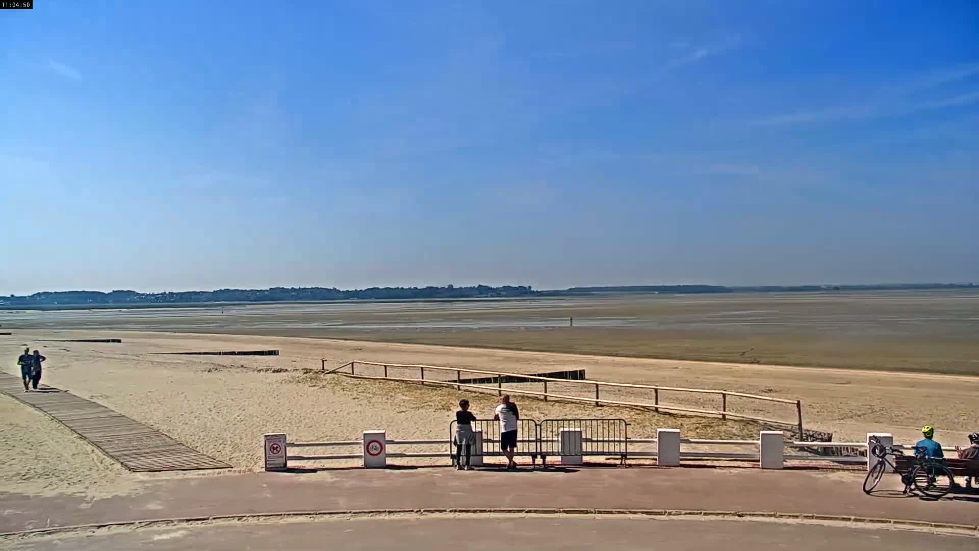 Under a clear blue sky, a sandy beach stretches toward a wide expanse of exposed mudflats, with several people relaxing on the beach and near a boardwalk.