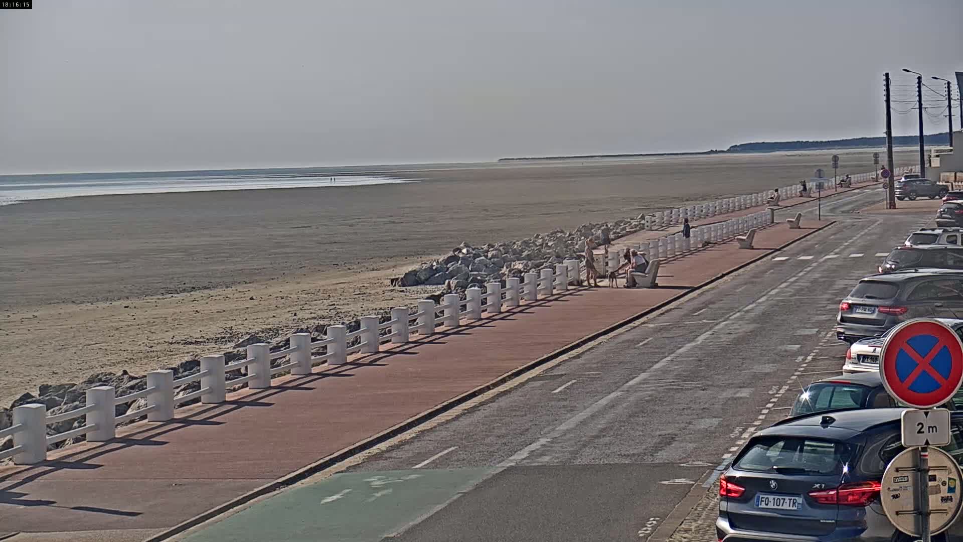 A wide sandy beach stretches to a calm sea under a hazy sky, alongside a paved walkway bordered by a white railing and lined with parked cars.