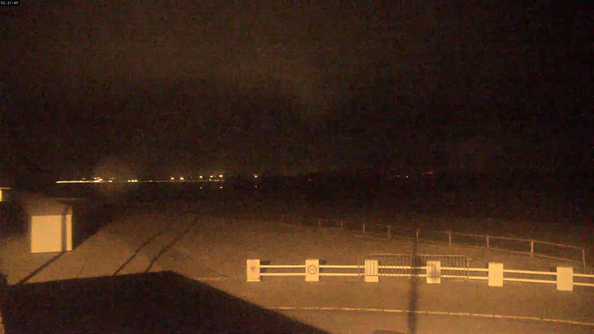 A nighttime view of a dark, flat area with a white fence and distant lights, possibly under a cloudy sky.