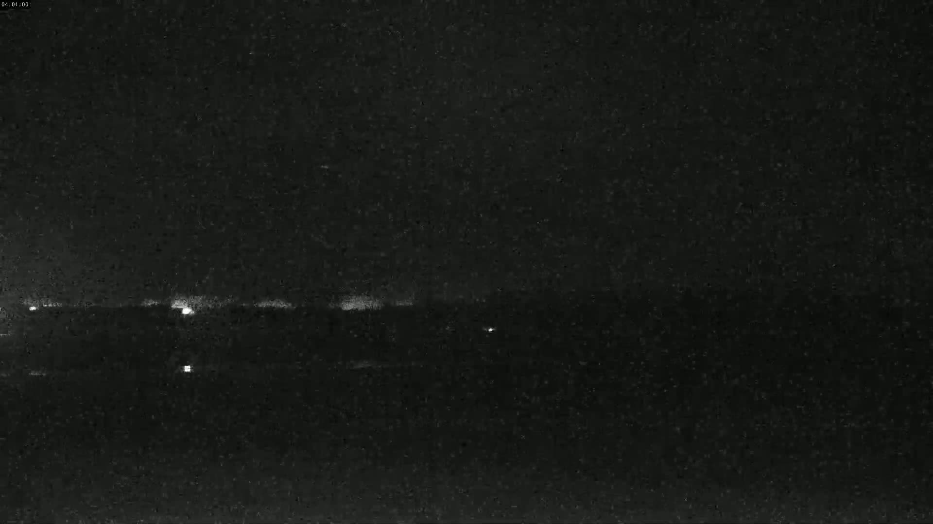 A nighttime, low-light image shows a dark landscape with a few faint lights in the distance under a seemingly clear, dark sky.