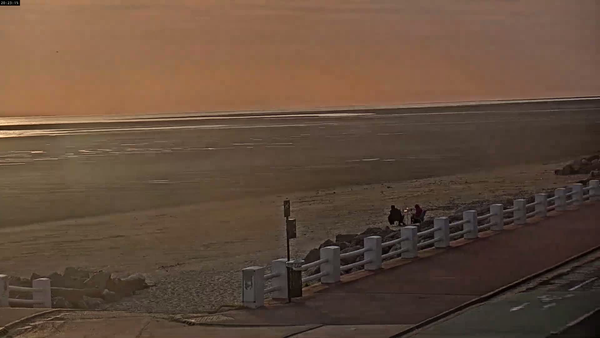 Two people sit on a sandy beach near a low white railing under an orange-toned sunset sky.
