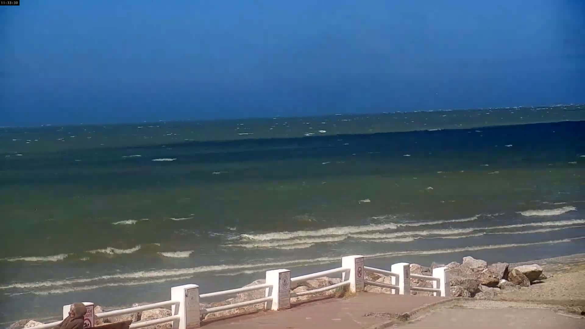 A mostly clear blue sky overlooks a wavy sea meeting a shoreline with a low white fence and rocks under a windy condition.