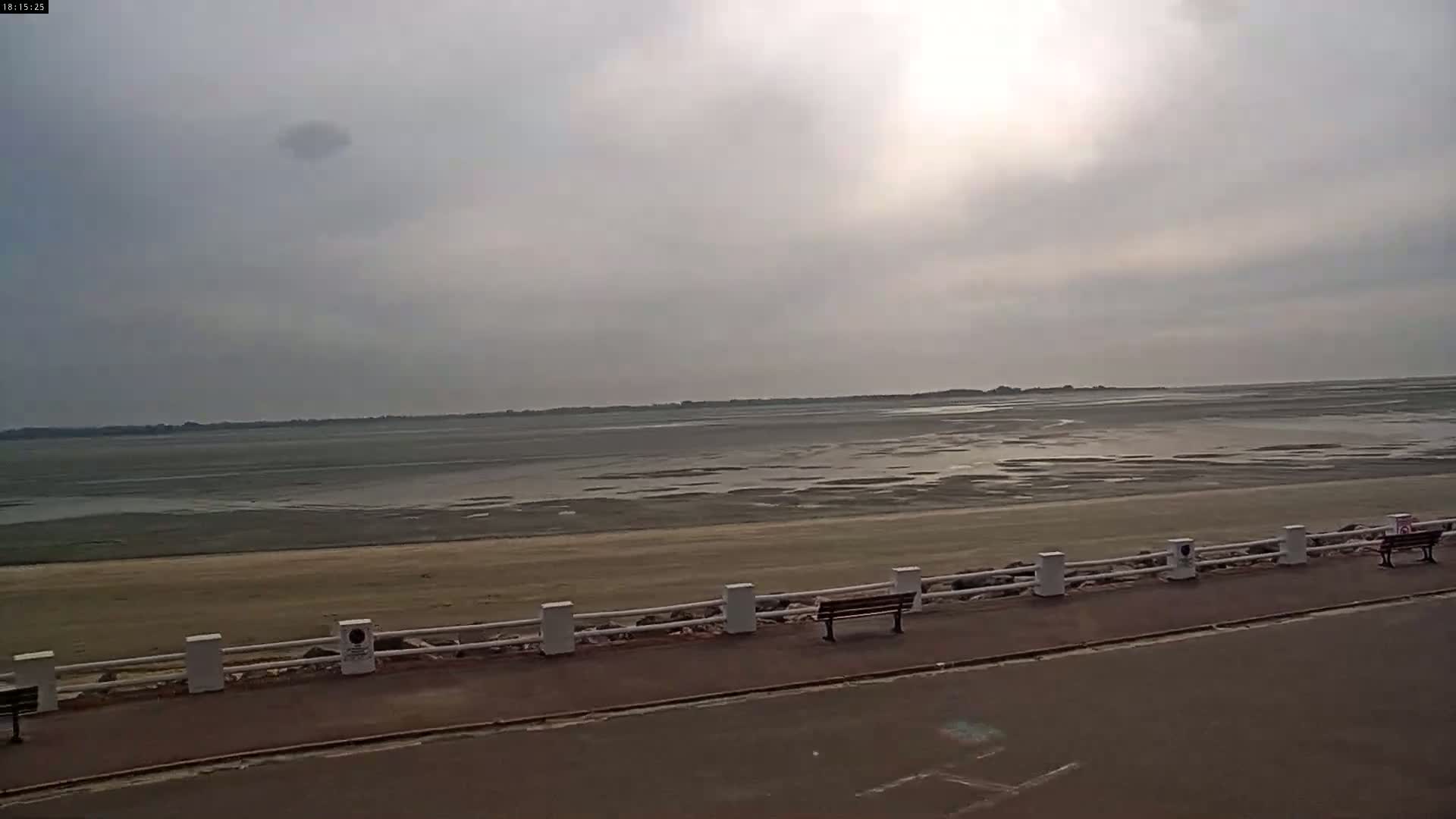 A mostly cloudy day reveals a wide expanse of mudflats at low tide, bordered by a paved walkway with a white fence and a bench.