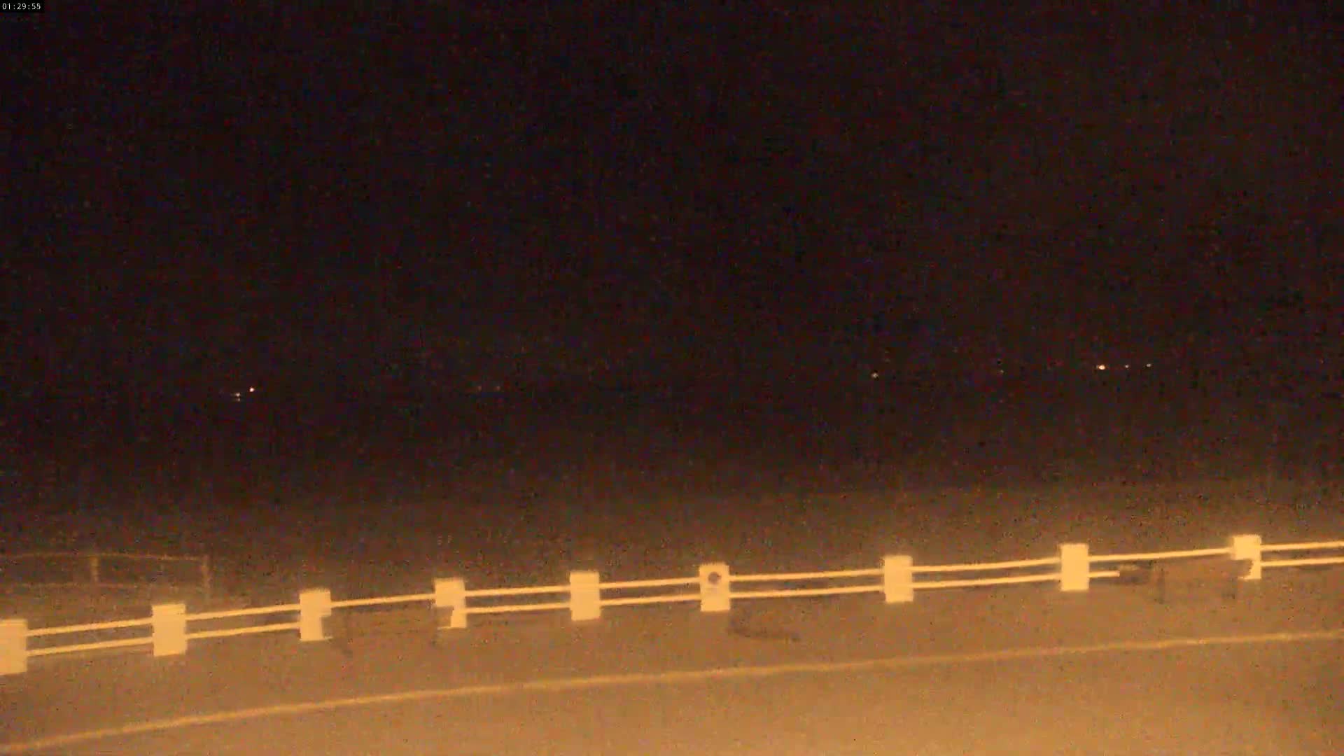 A nighttime view of a road with a low white fence, under a dark sky with a few distant lights visible.