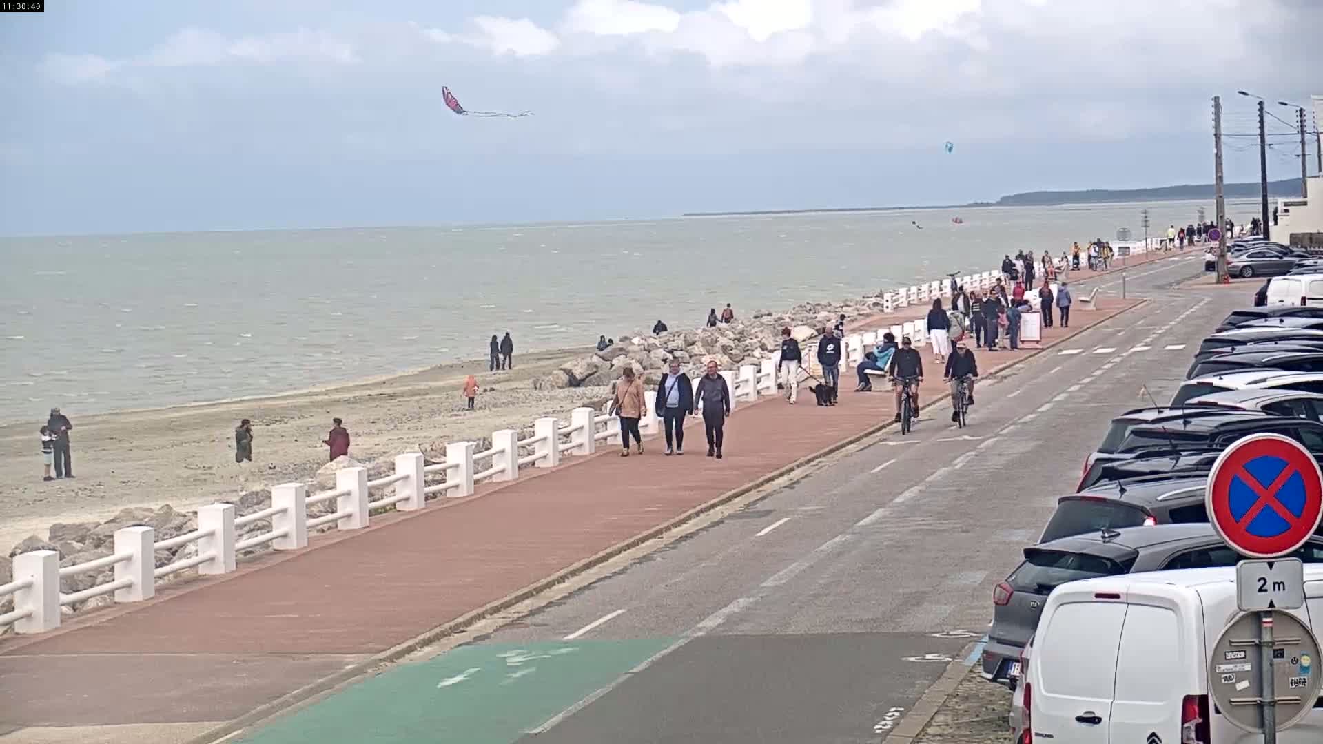 A paved walkway beside a calm sea, with people walking, biking, and flying kites on a partly cloudy day.