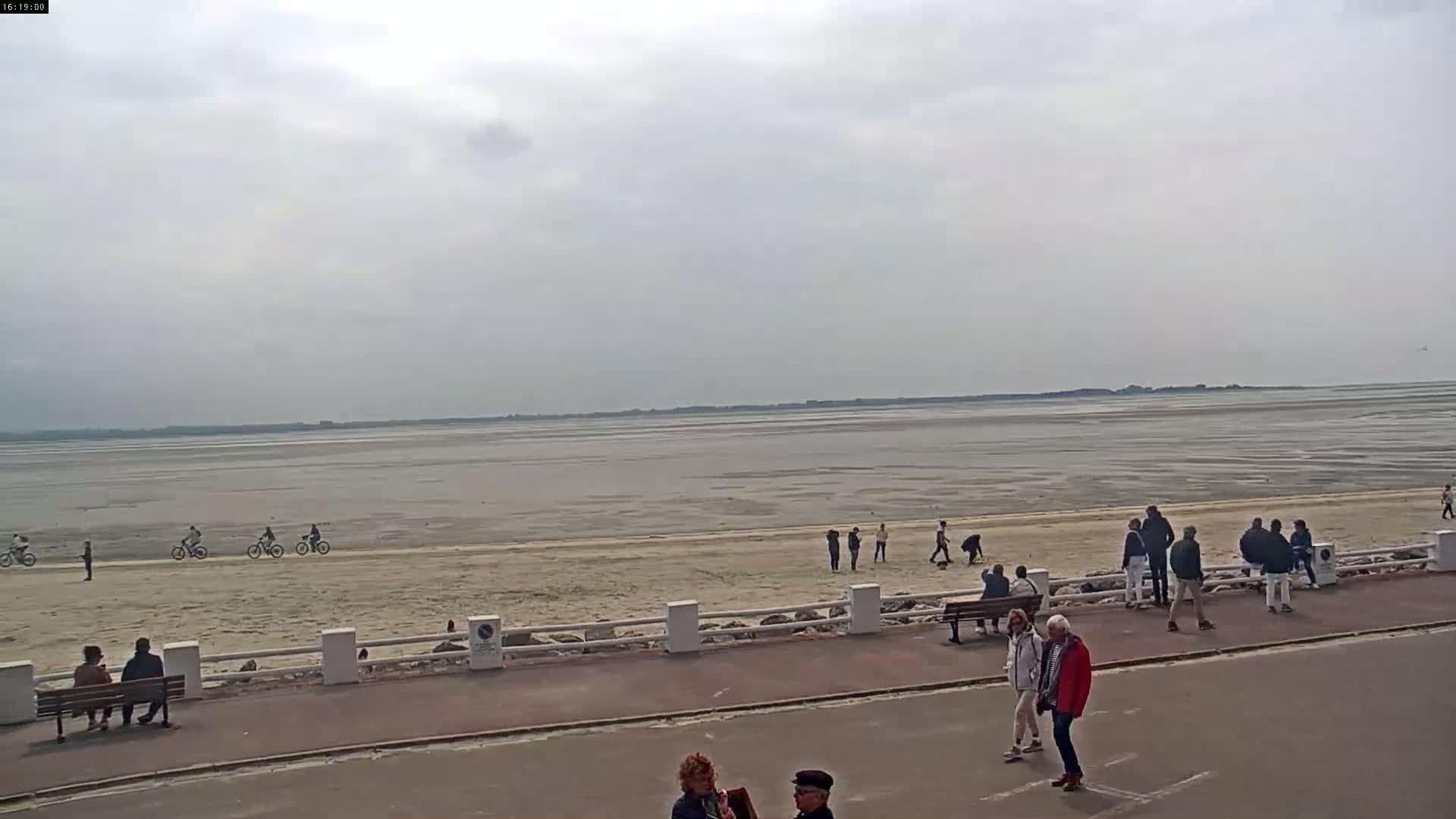A mostly overcast day shows a wide expanse of exposed sand and shallow water, with people walking and riding bicycles on the beach and along a paved walkway beside a low white wall.