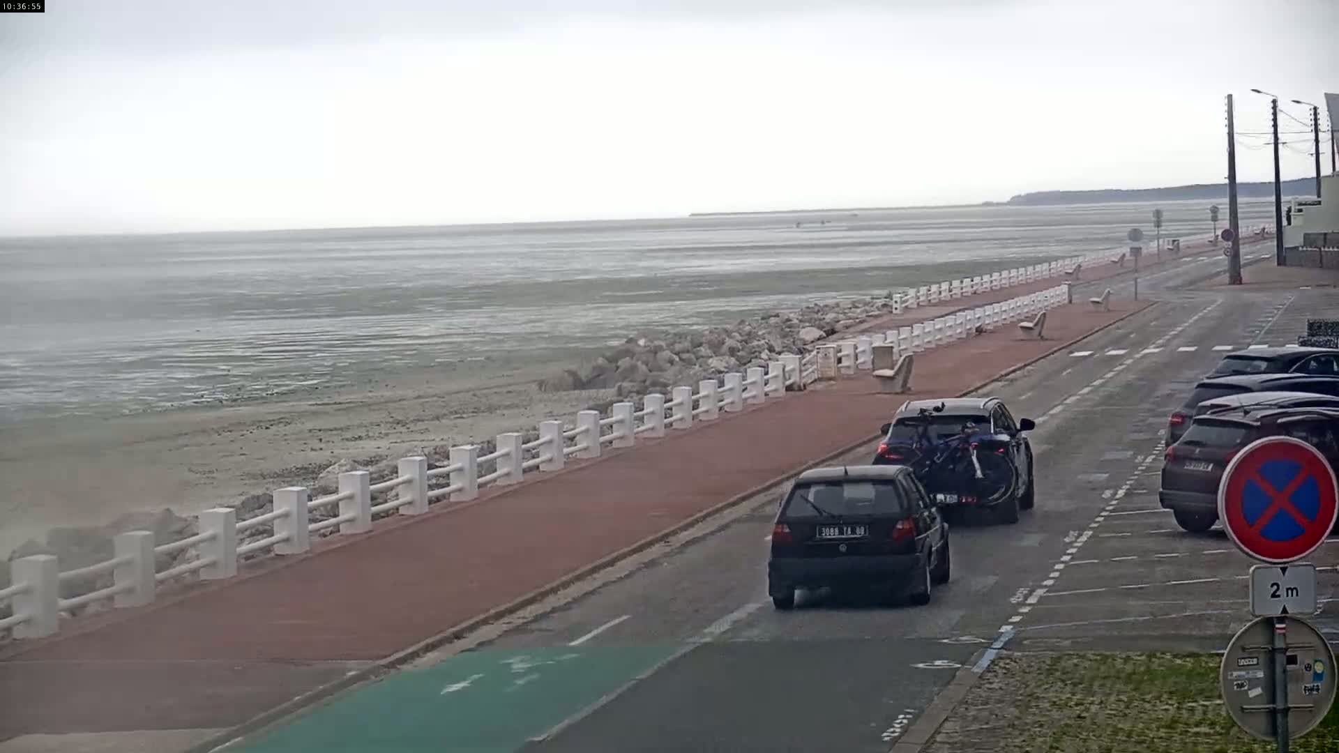 A coastal road with parked cars runs alongside a seawall bordering a calm, grey sea under an overcast sky.