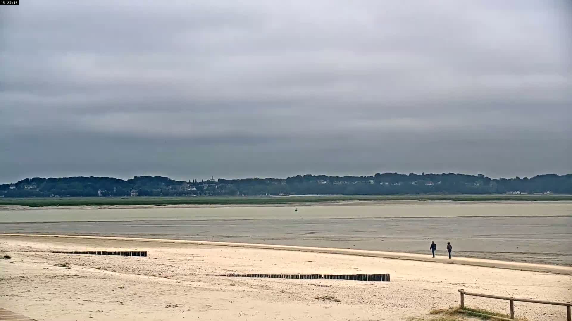 A sandy beach stretches towards a calm, pale-colored bay under an overcast sky, with two people walking on the shore in the distance.