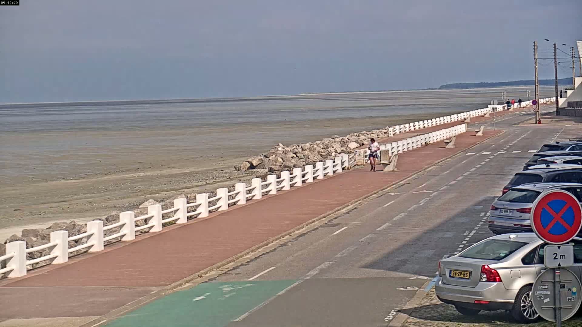 A person walks along a paved path next to a low white fence separating the path from a rocky shore and a wide expanse of exposed tidal flats under a mostly clear sky.