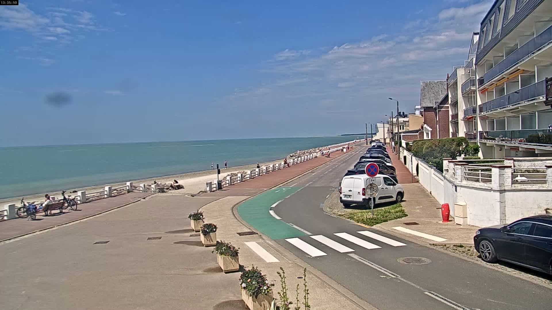 A seaside promenade with parked cars and a few pedestrians, under a mostly sunny sky, leads to a calm, light-blue ocean.