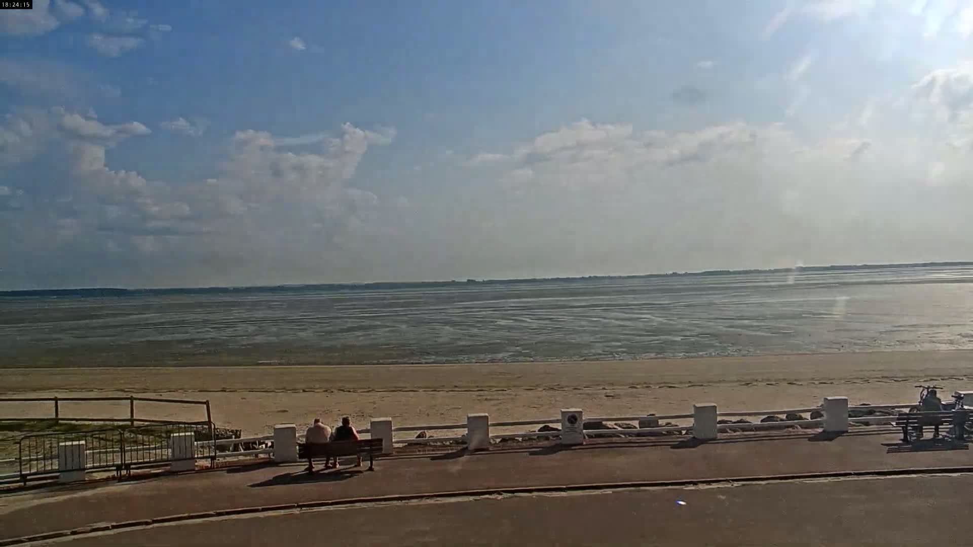 A partly cloudy day reveals a wide expanse of sandy beach and calm, shallow water receding from the shore, where several people sit on benches along a paved walkway.