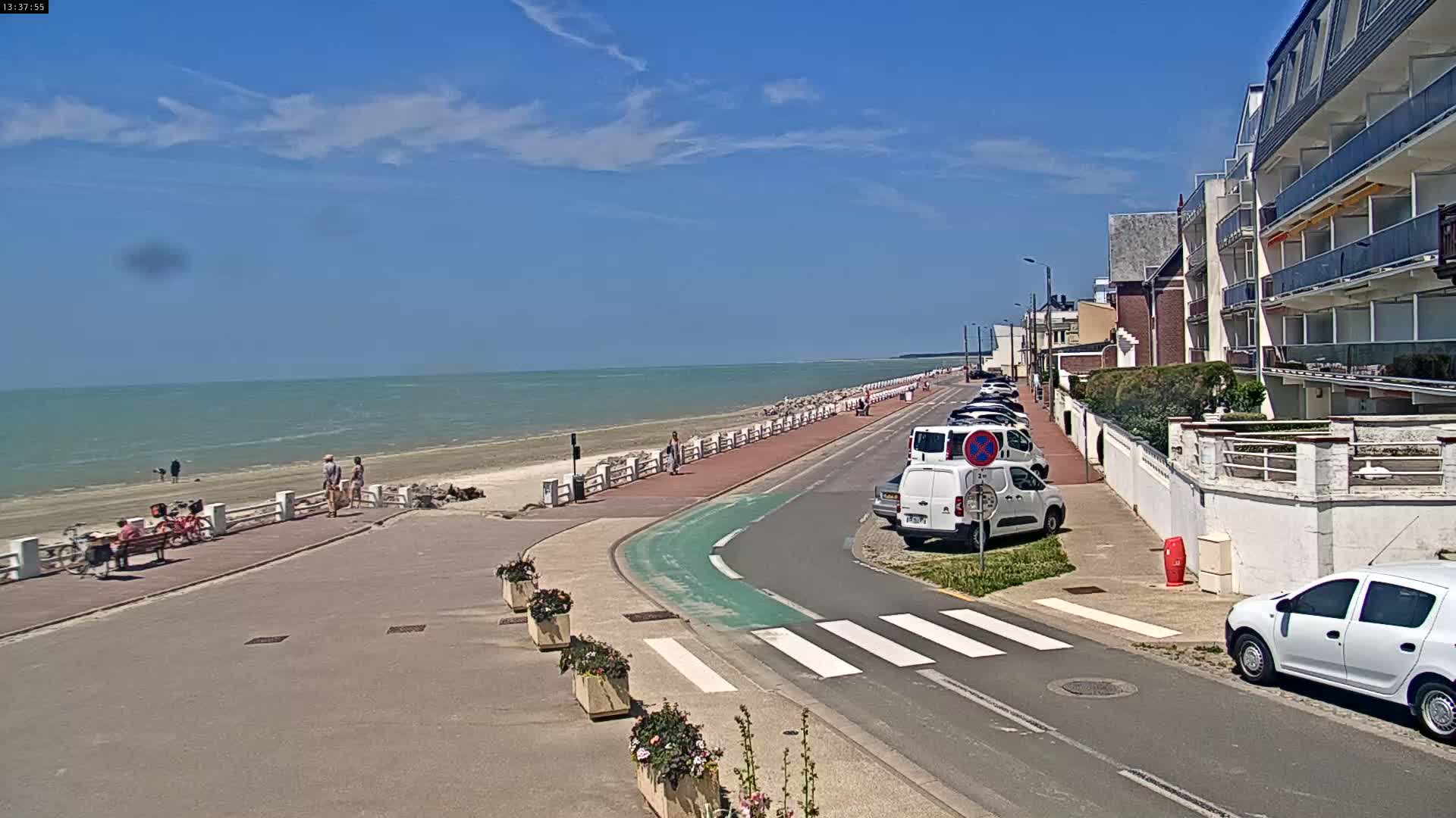 A sunny day at a seaside promenade shows parked cars along a road next to a beach with people walking along the walkway and on the sand.