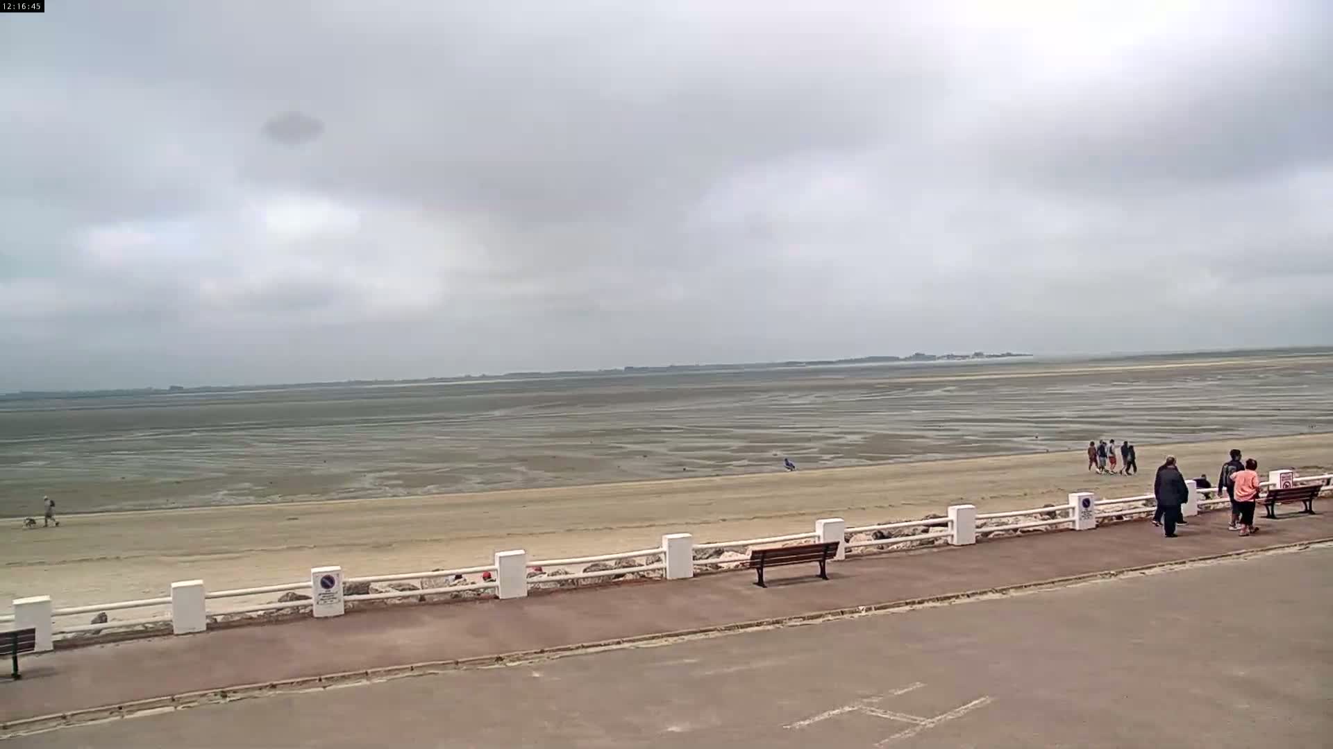 A wide, sandy beach stretches to a calm, gray-cloudy sky, with several people walking along the shore and sitting on benches near a low white railing.