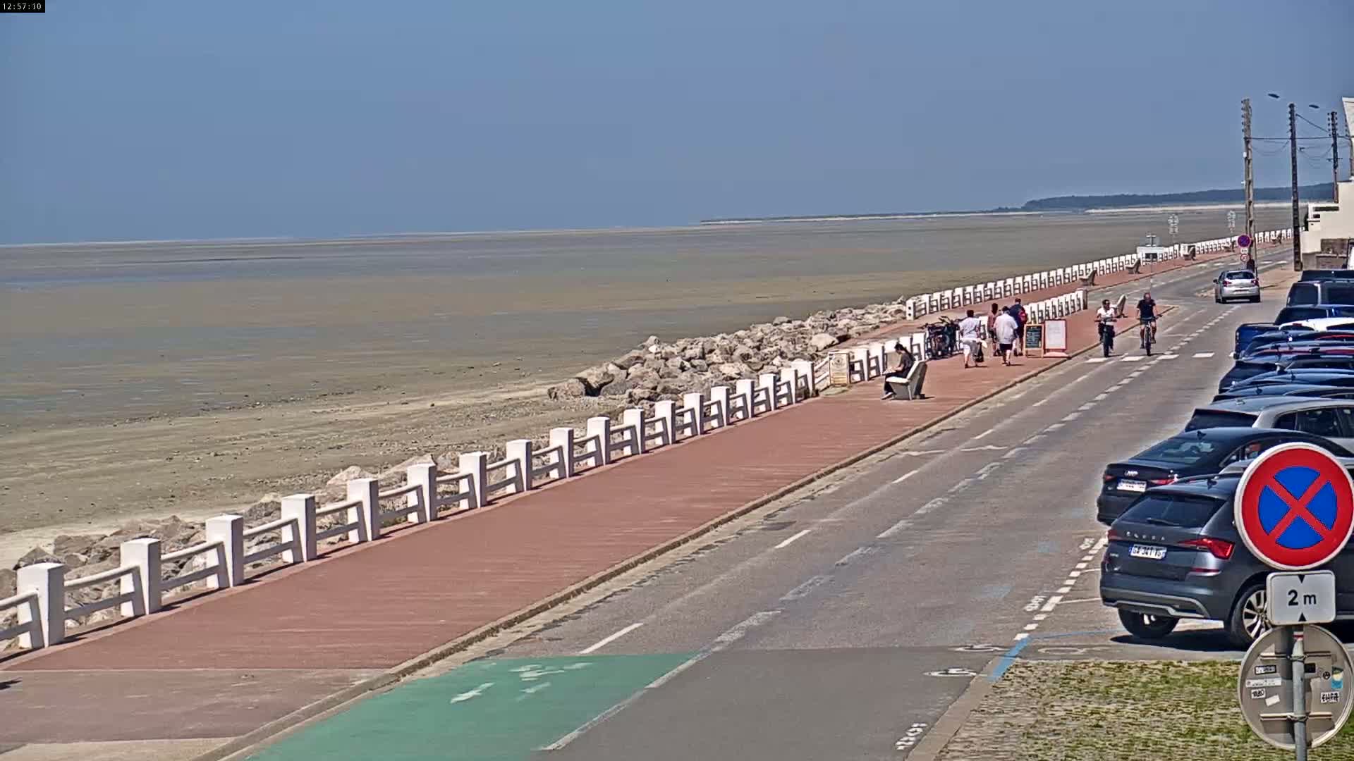 A paved walkway with a white railing lines a beach with exposed mudflats under a clear blue sky, and cars are parked along a road adjacent to the walkway.