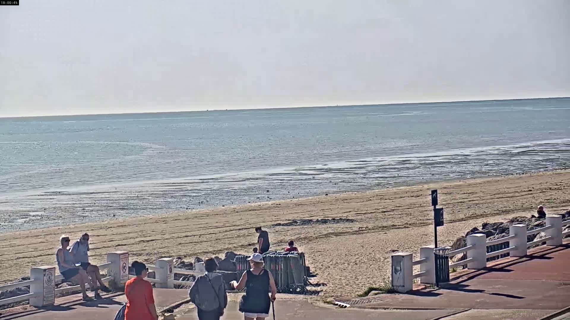 A sandy beach with several people on it and near a walkway, overlooks a calm sea under a clear sky.