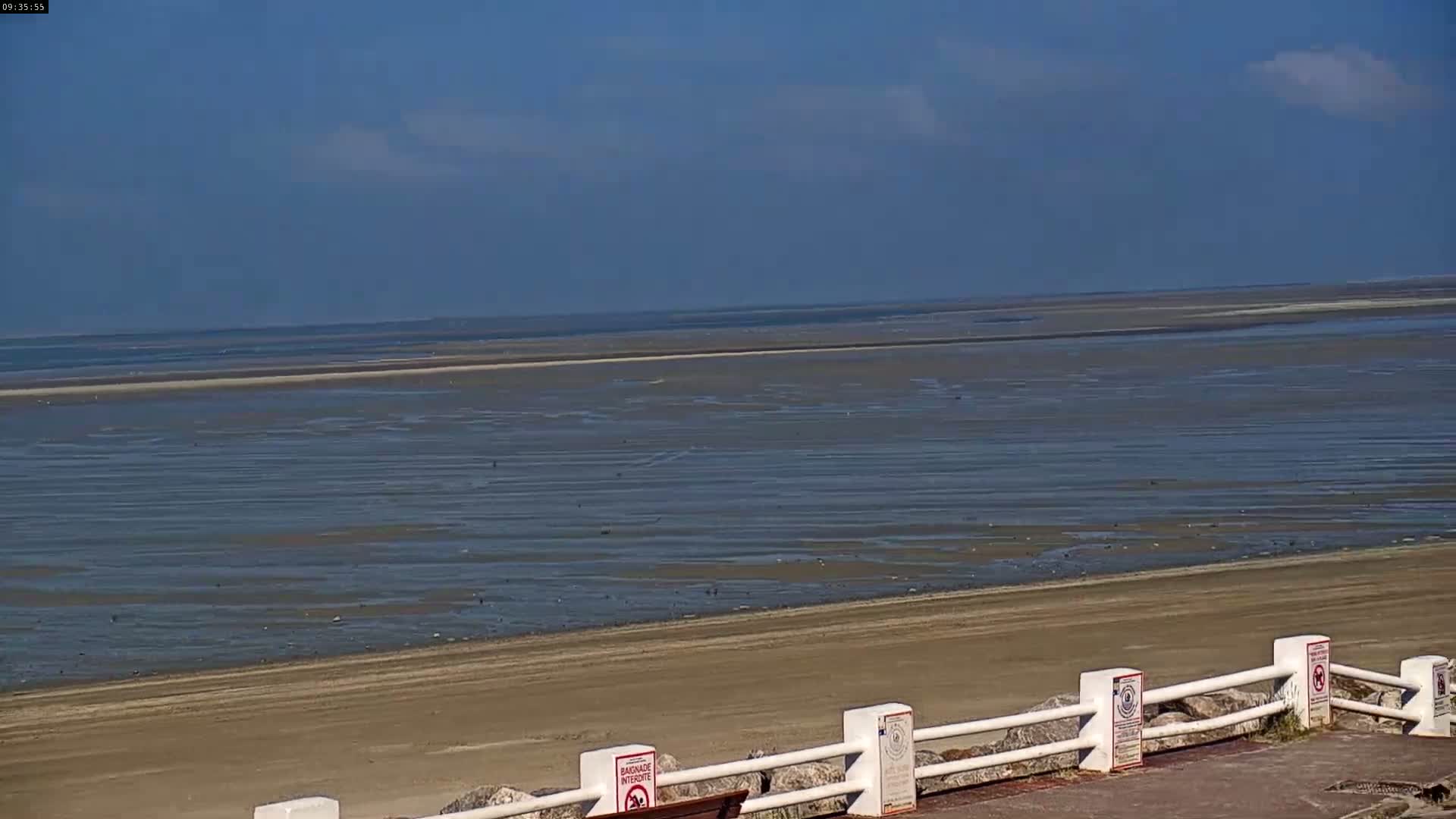 A mostly sunny day reveals a wide expanse of sandy beach and shallow, receding ocean water, bordered by a low white fence.