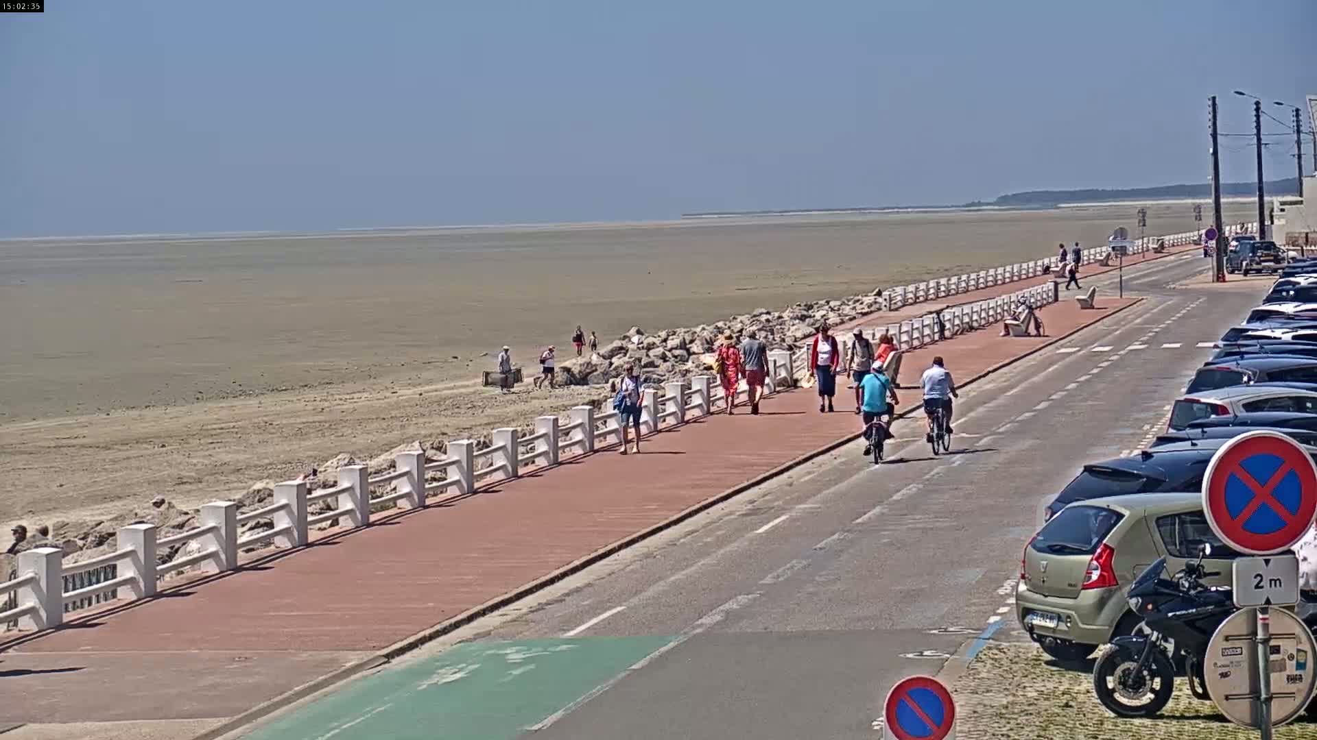 A paved walkway and road run alongside a beach with low tide, where several people are walking and cycling on a sunny day.