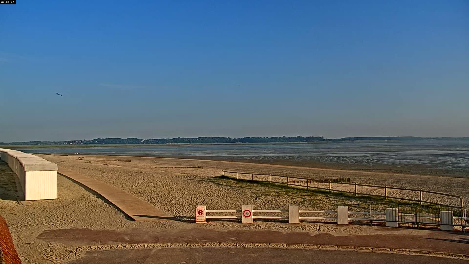 A sandy beach with white beach huts and a wooden walkway stretches towards a calm, low-tide bay under a clear blue sky.