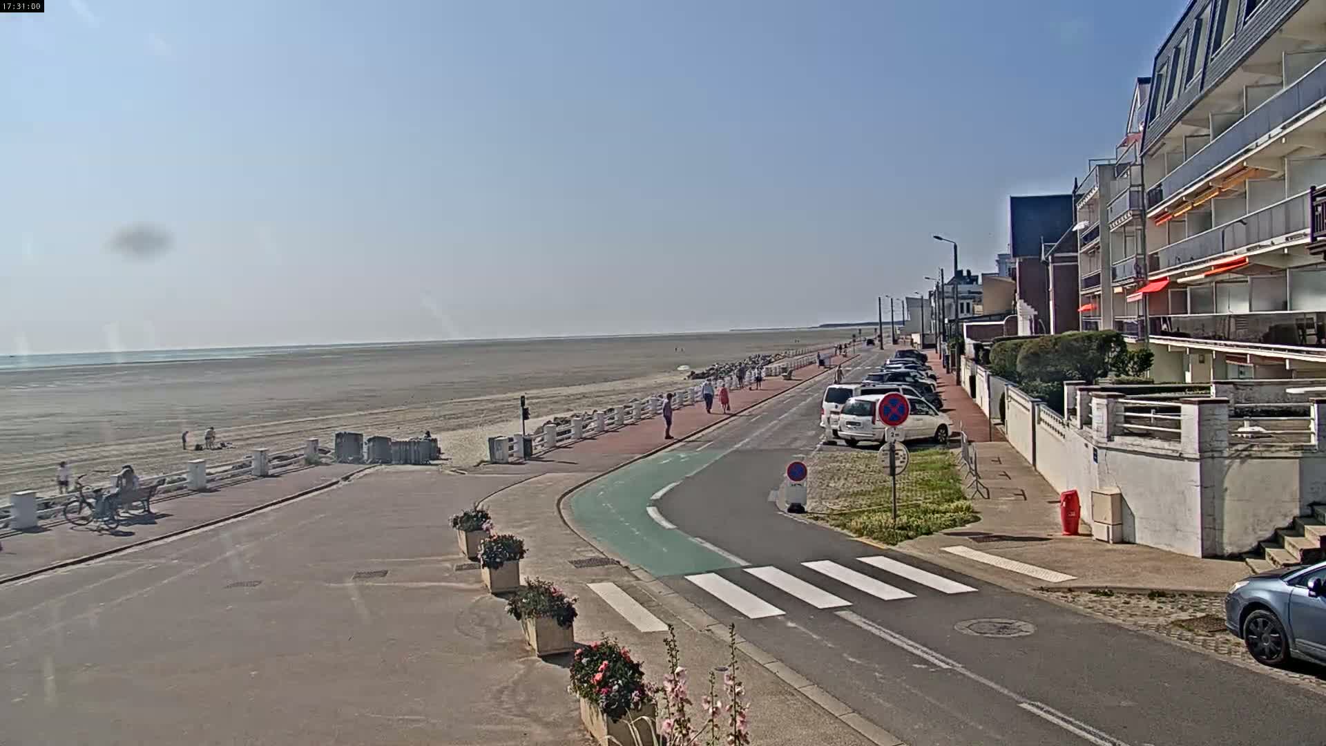 A wide shot shows a beach with a few people walking on it, next to a paved road with parked cars and buildings, under a clear sky.