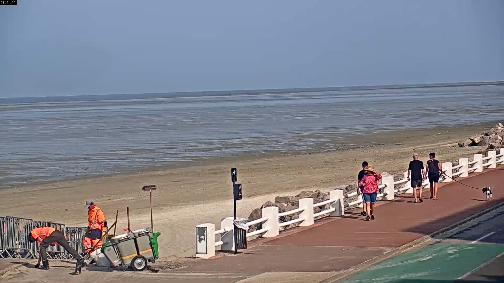 On a sunny day, two sanitation workers clean a beach while several people stroll along a paved walkway beside a calm, shallow body of water.