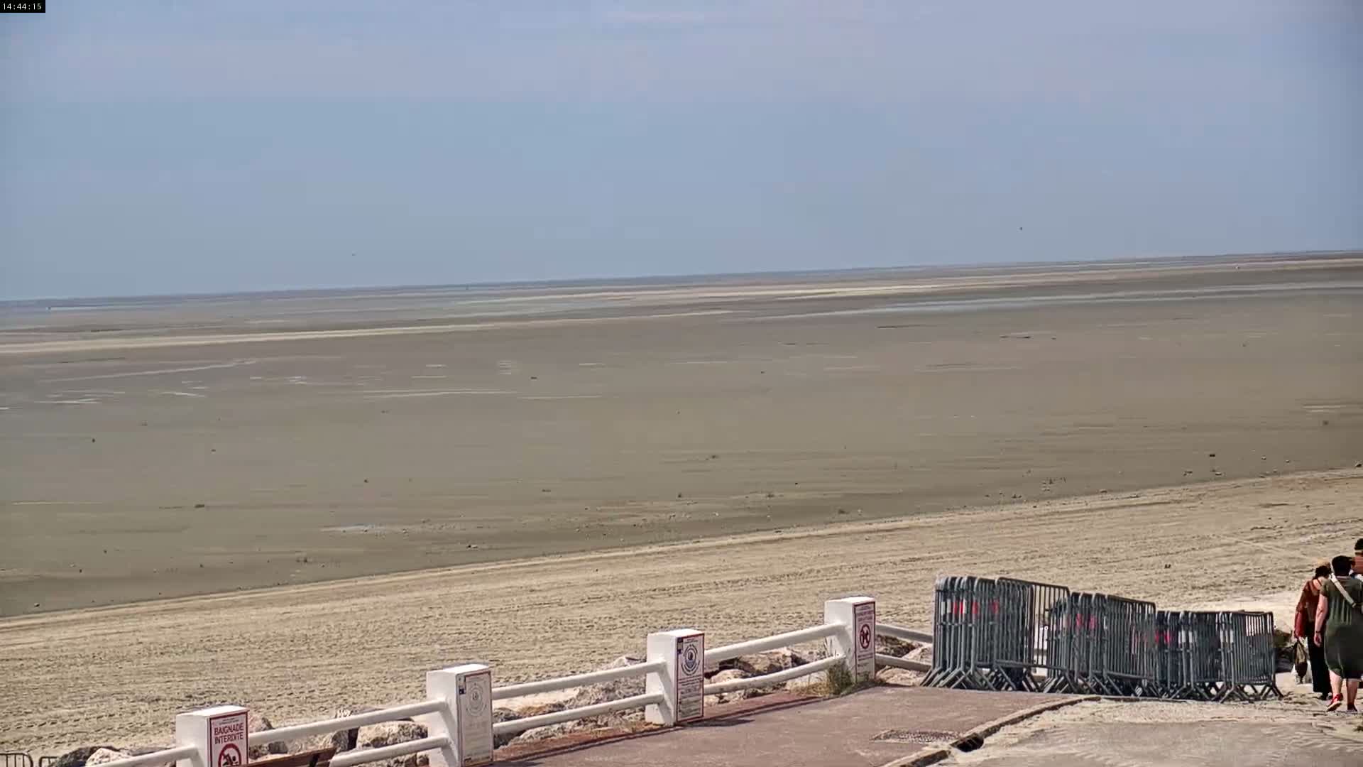 A wide expanse of flat, light-brown sand extends to a calm, light-blue ocean under a clear sky, with a few people walking near a low white railing on the shore.