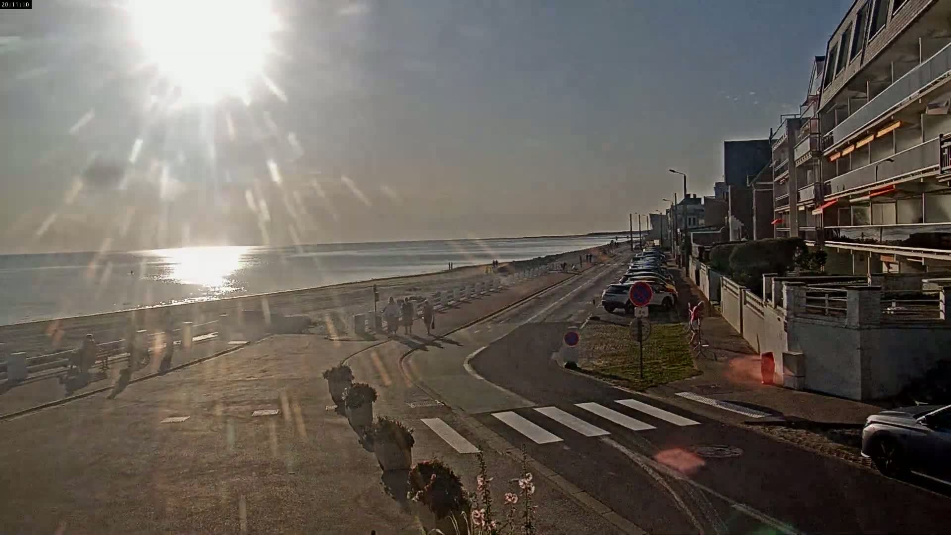 A sunny day at a beachside promenade shows pedestrians strolling along a paved path next to a calm ocean, with buildings and parked cars lining the street.