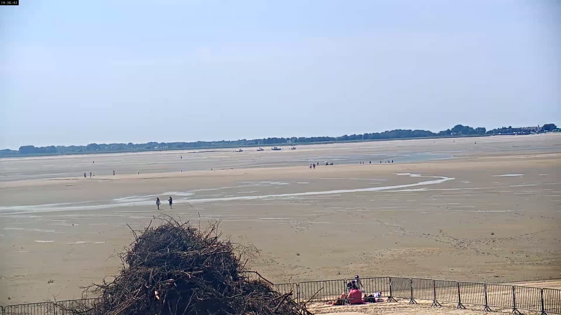 A wide expanse of flat, sandy beach stretches to a distant shoreline under a clear, sunny sky, with a few people scattered across the beach and several boats visible in the water.