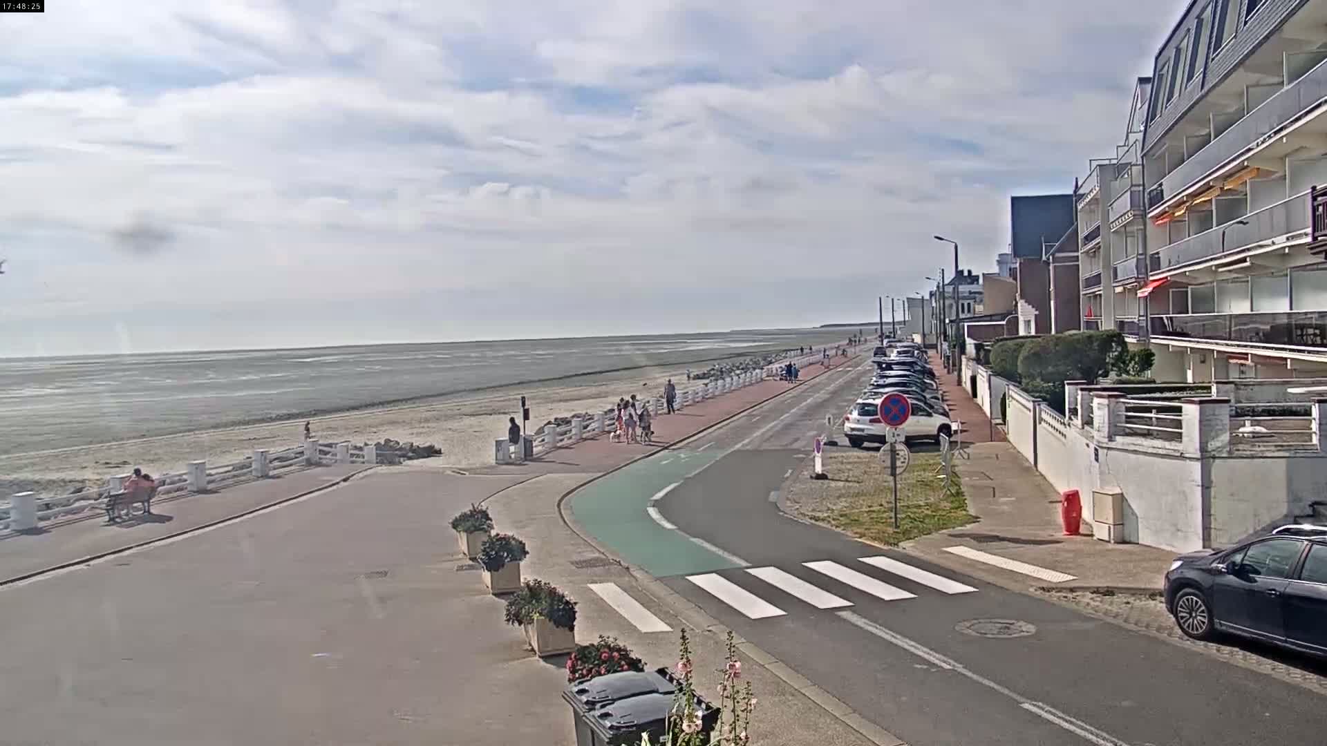 A paved road with a pedestrian crossing leads to a beach with people walking along the shoreline under a partly cloudy sky.