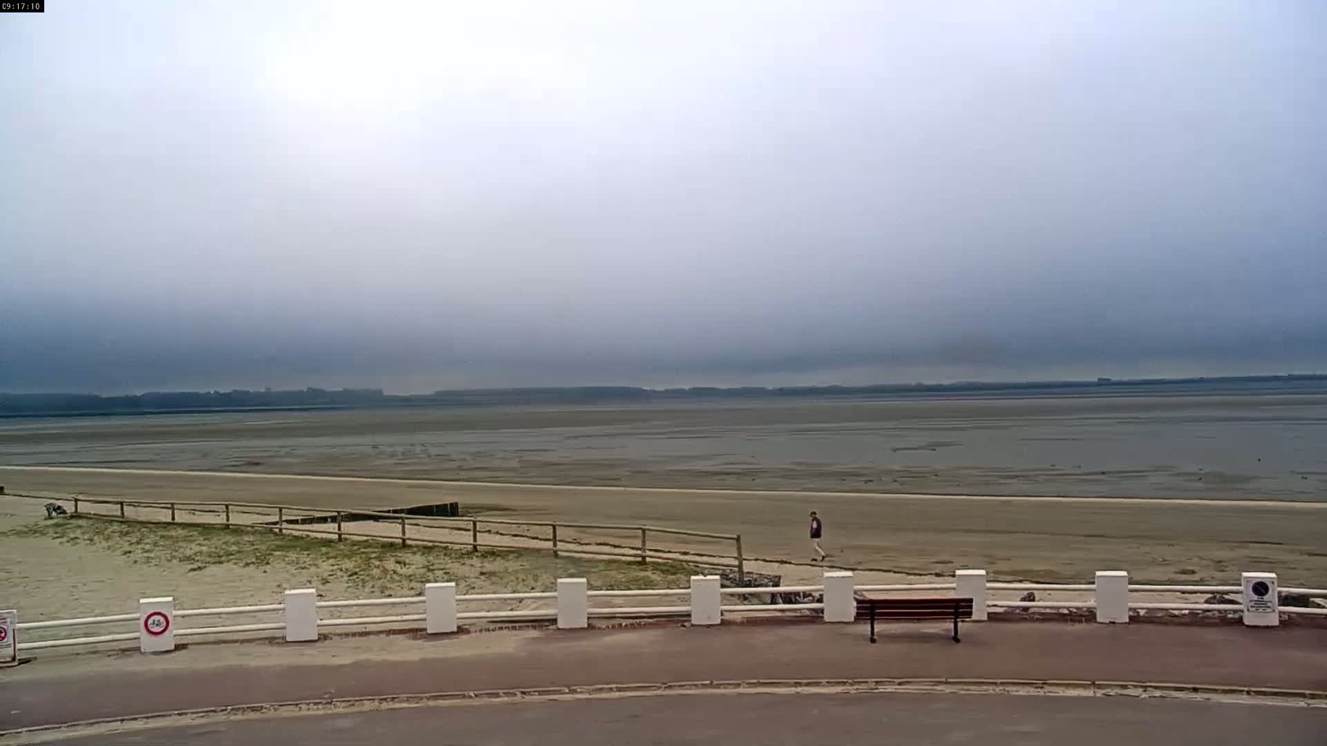 A person walks along a sandy beach under an overcast sky, past a wooden bench and white fence.