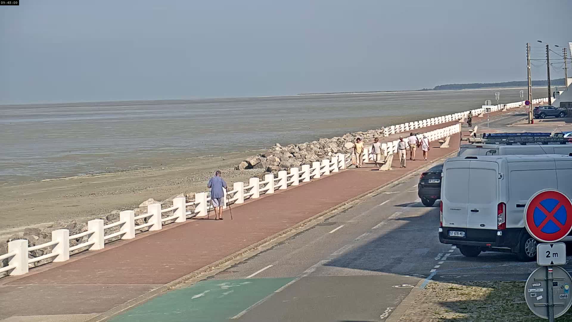 A paved walkway runs alongside a calm, wide body of shallow, muddy water, where several people are strolling, and cars are parked along the roadside.