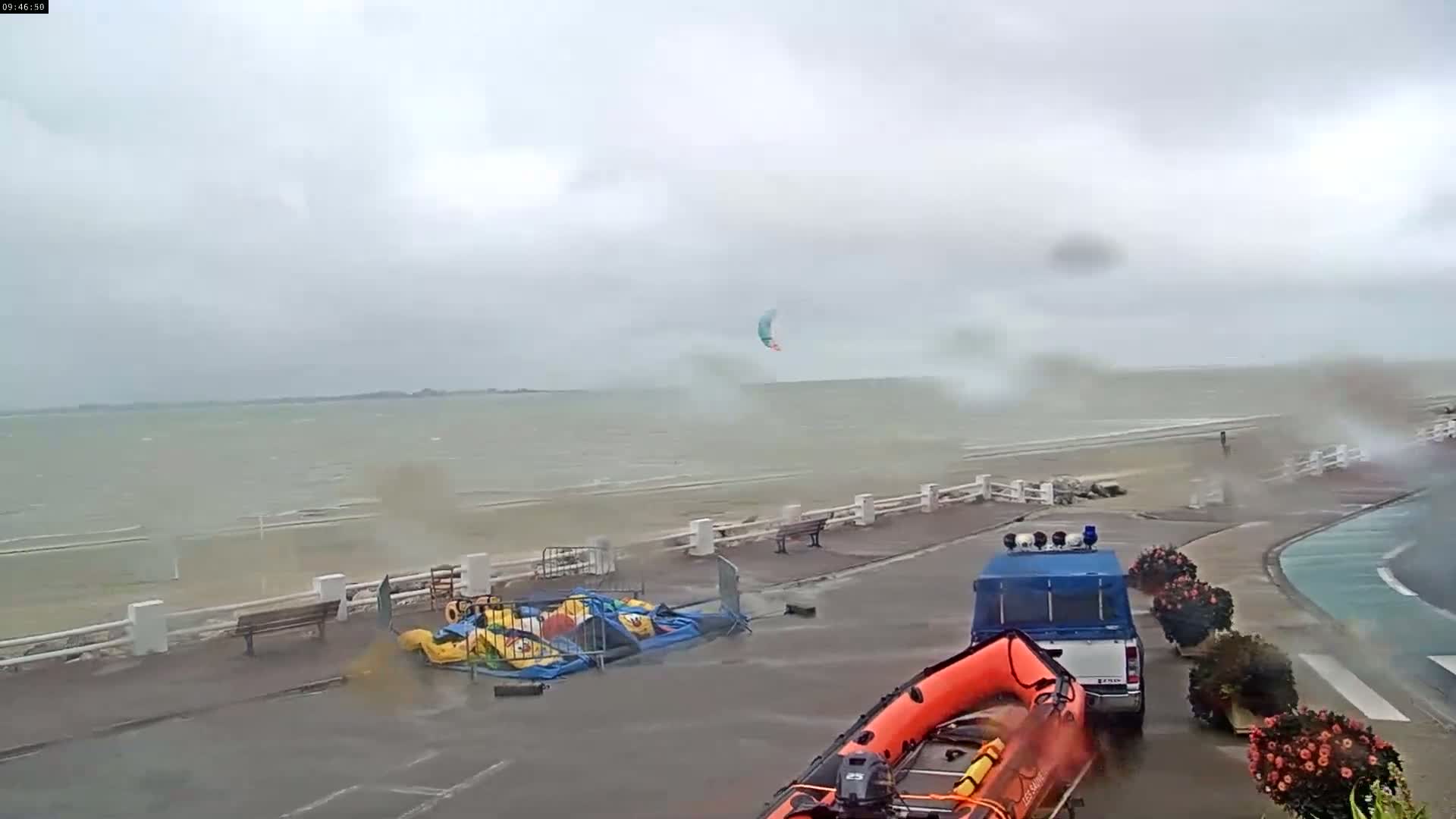 A windy, overcast day at the beach shows a kitesurfer in the distance, a lifeboat, and a truck parked near some blown-over inflatable playground equipment.