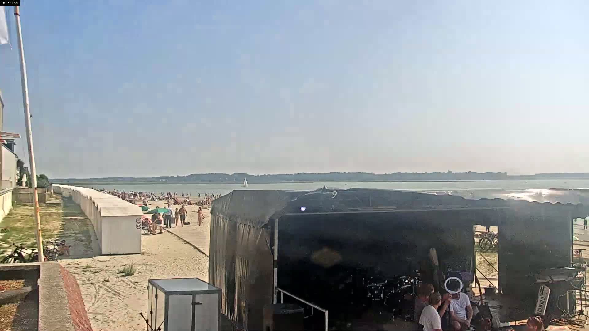 A wide expanse of muddy tidal flats stretches to a calm sea under a mostly cloudy sky, viewed from behind a low white railing on a sandy beach.