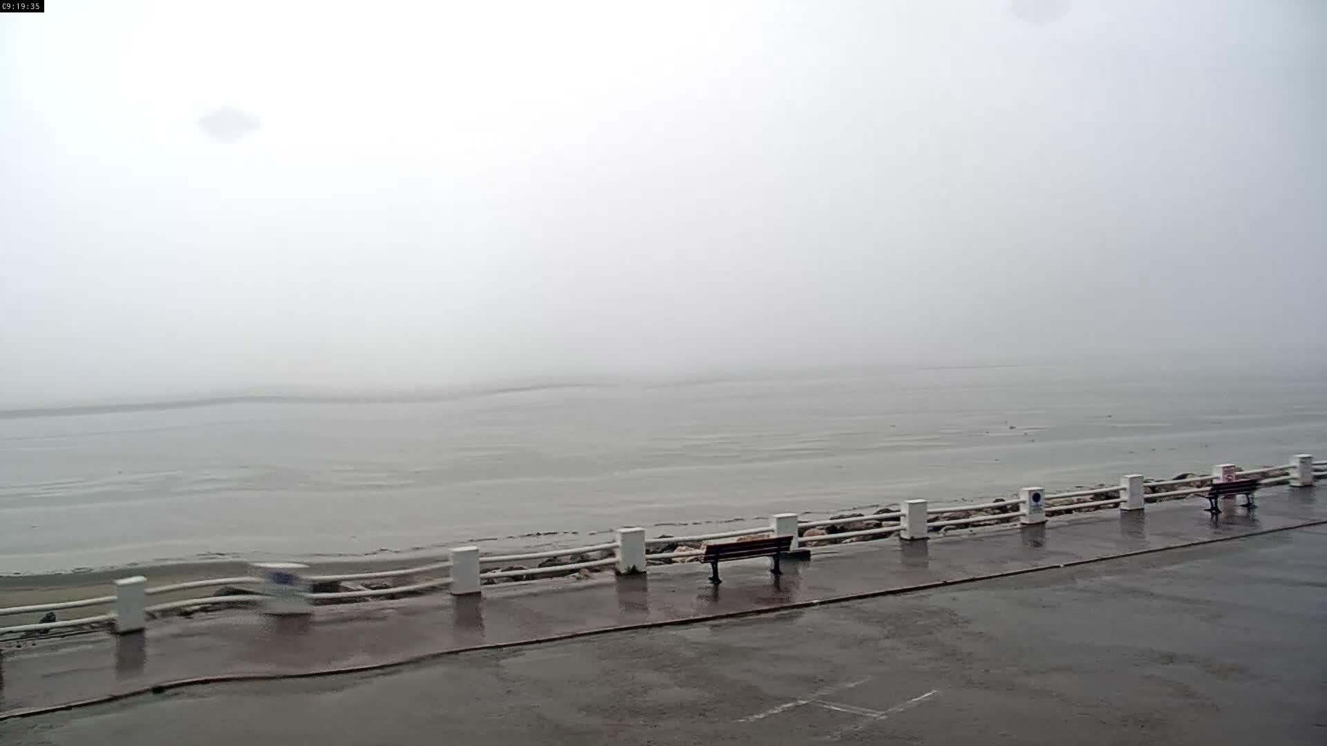 A wide expanse of muddy tidal flats stretches to a calm sea under a mostly cloudy sky, viewed from behind a low white railing on a sandy beach.