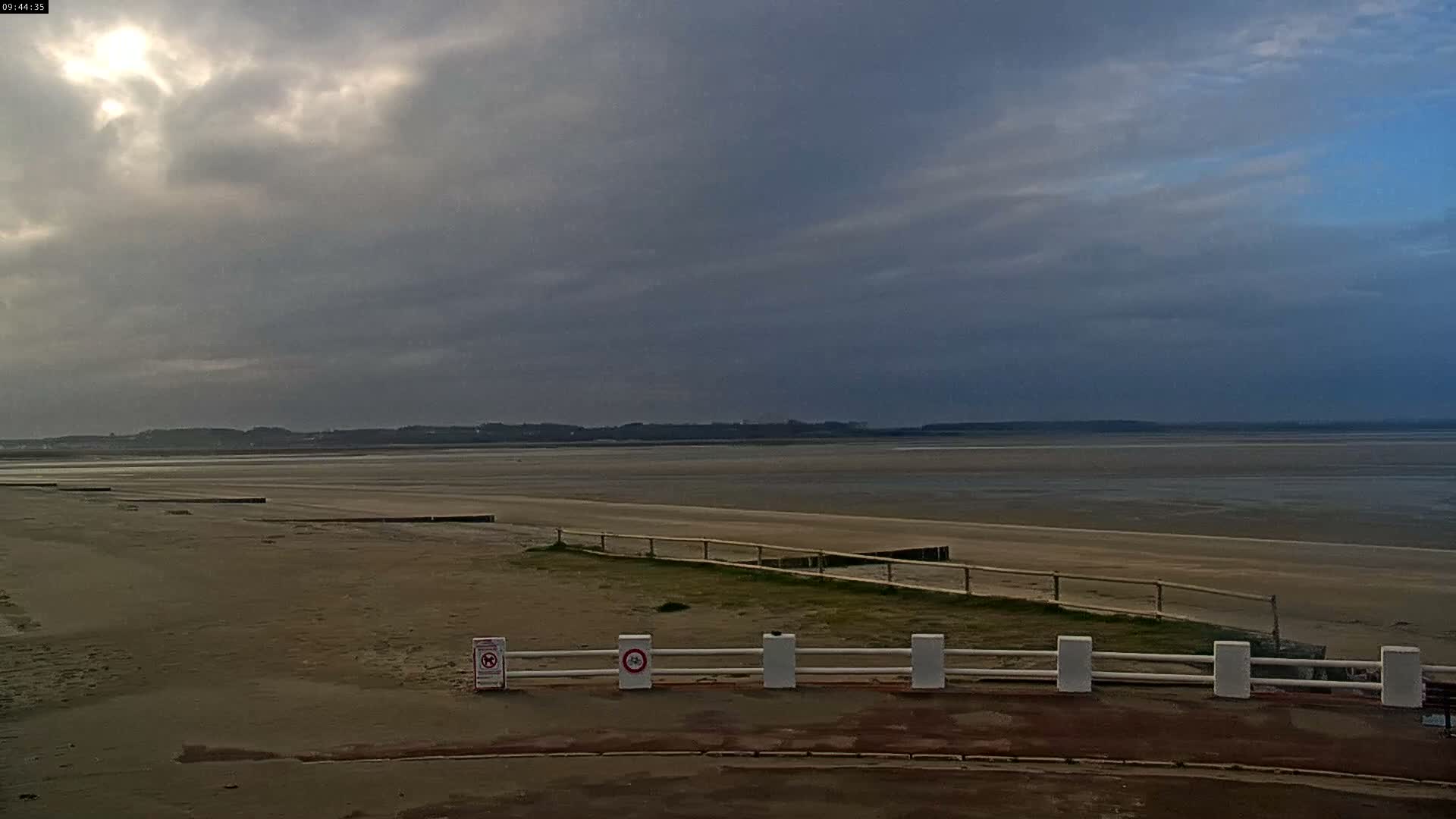 An expansive sandy tidal flat stretches towards a distant coastline under a heavily overcast sky where patches of light indicate the sun attempting to break through.